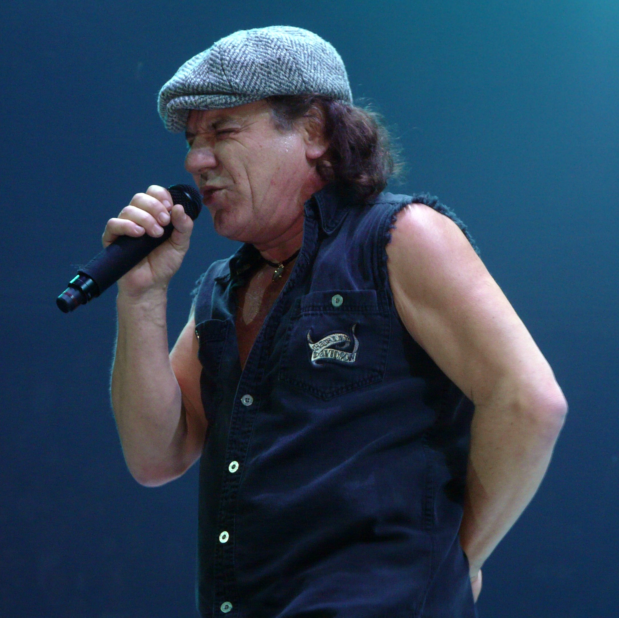 Brian Johnson singing on stage, wearing his signature flat cap and holding a microphone
