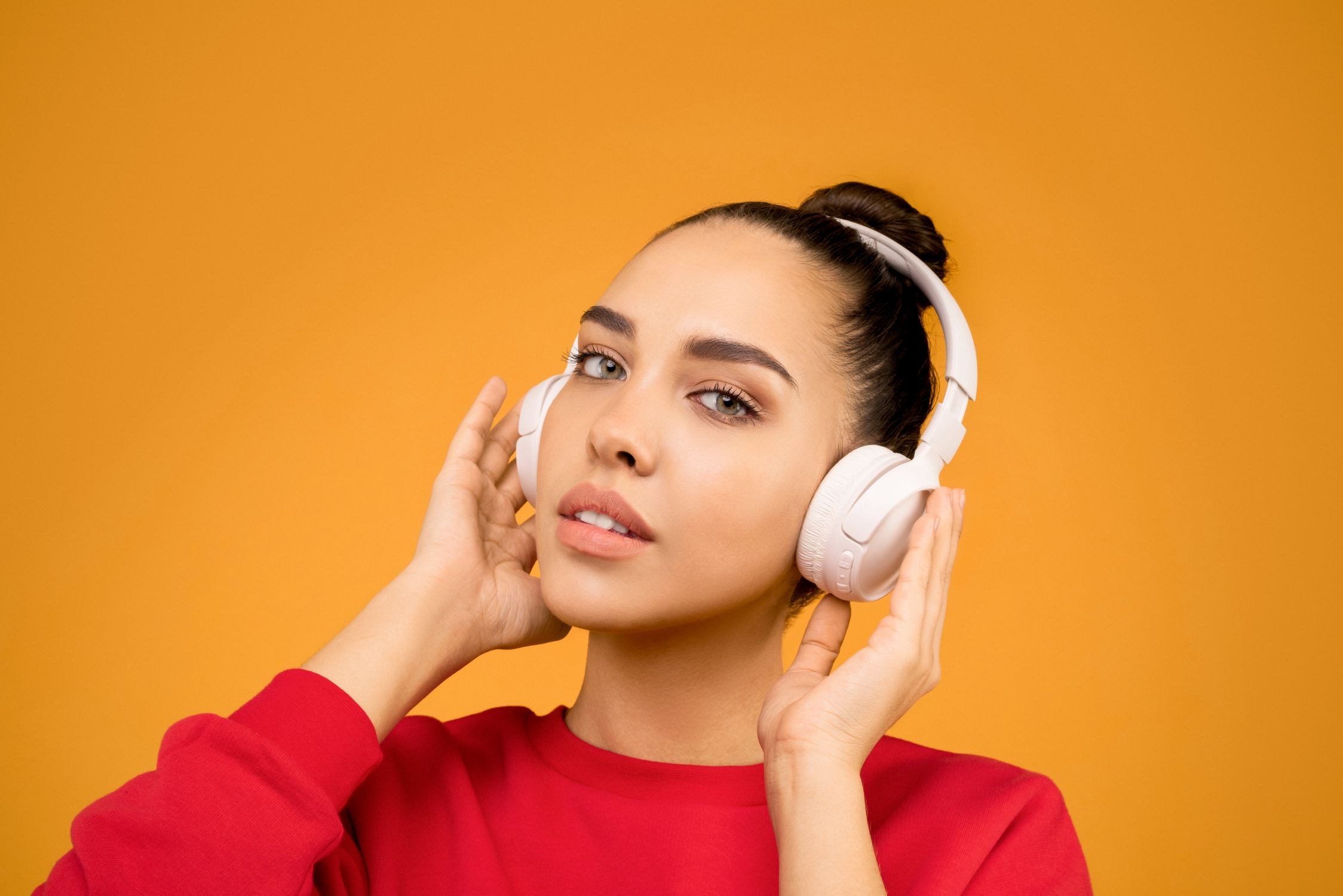 A young woman listening with white headphones against a bright orange background