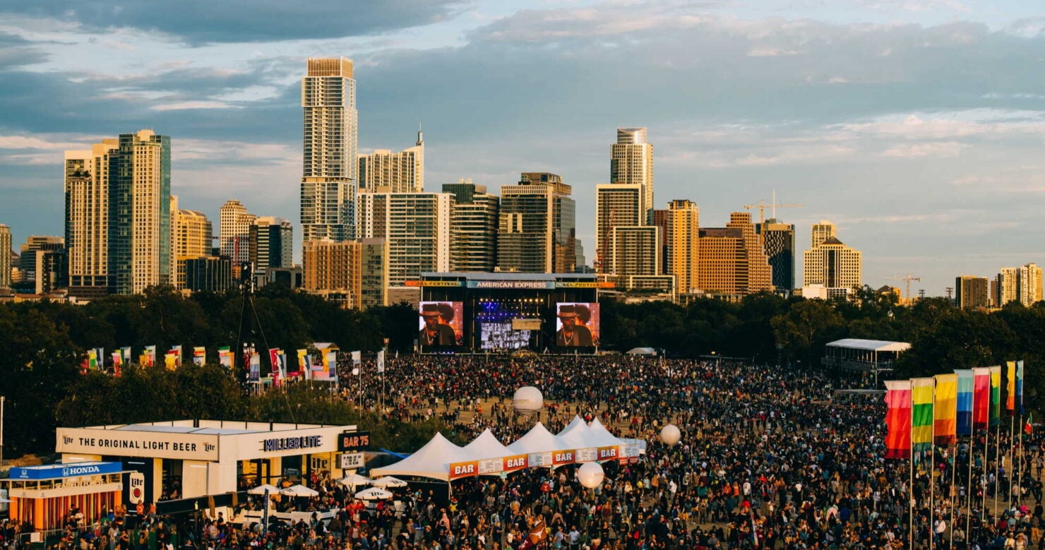 A crowd gathers in front of the American Express stage at Austin City Limits in Zilker Park, the downtown Austin skyline behind