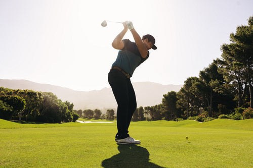 A golfer swinging in bright daylight on a scenic championship golf course.