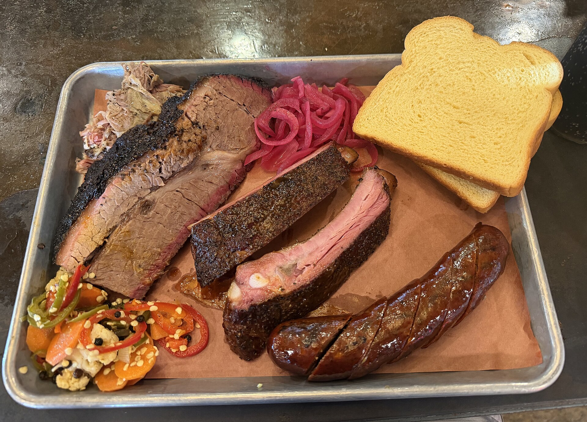 A Texas-style barbecue tray with sliced brisket, pork ribs, sausage, pulled pork, white bread, and pickled onions