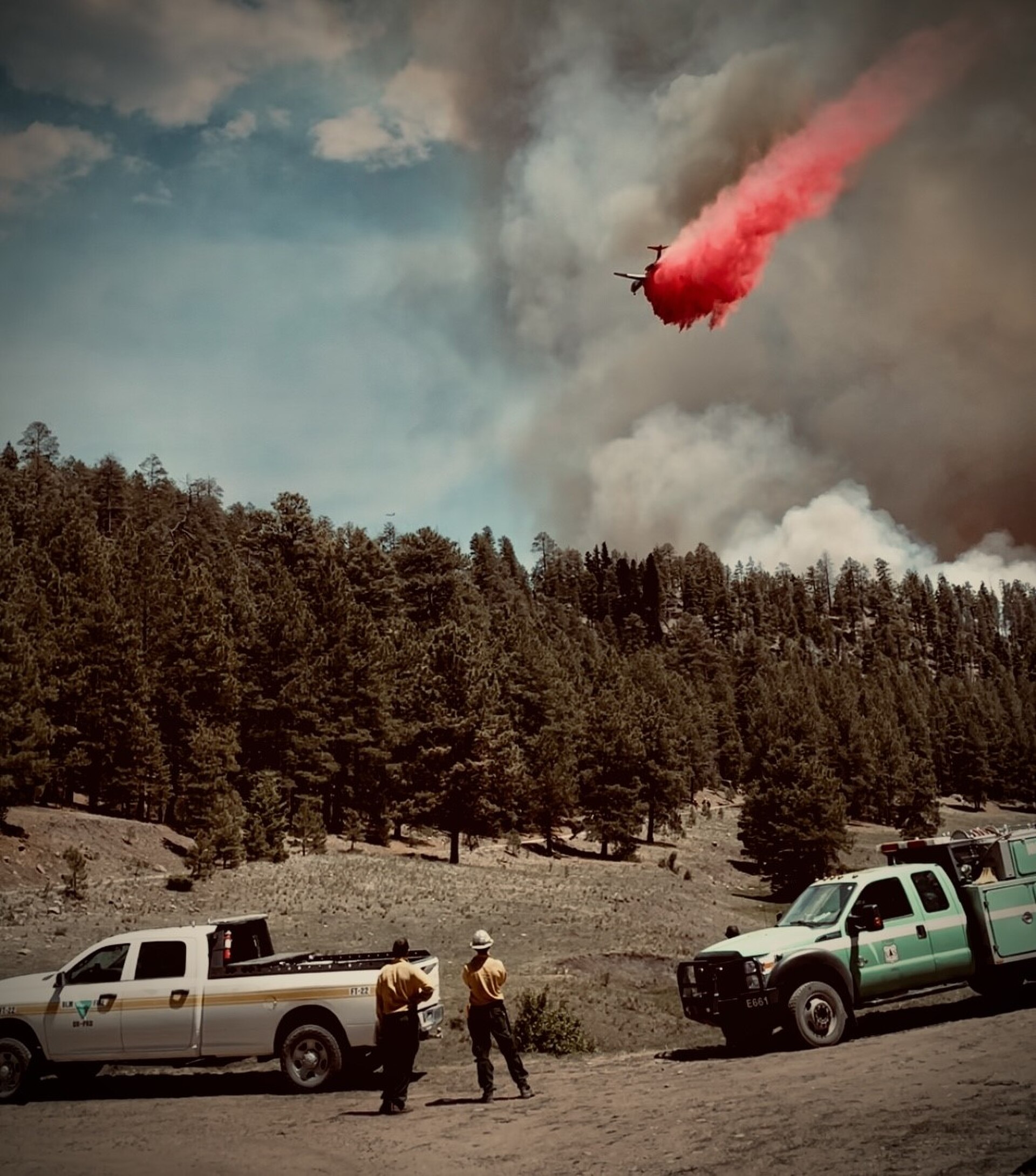 An air tanker drops bright-red fire retardant over a forested ridgeline in daylight while firefighters watch from below