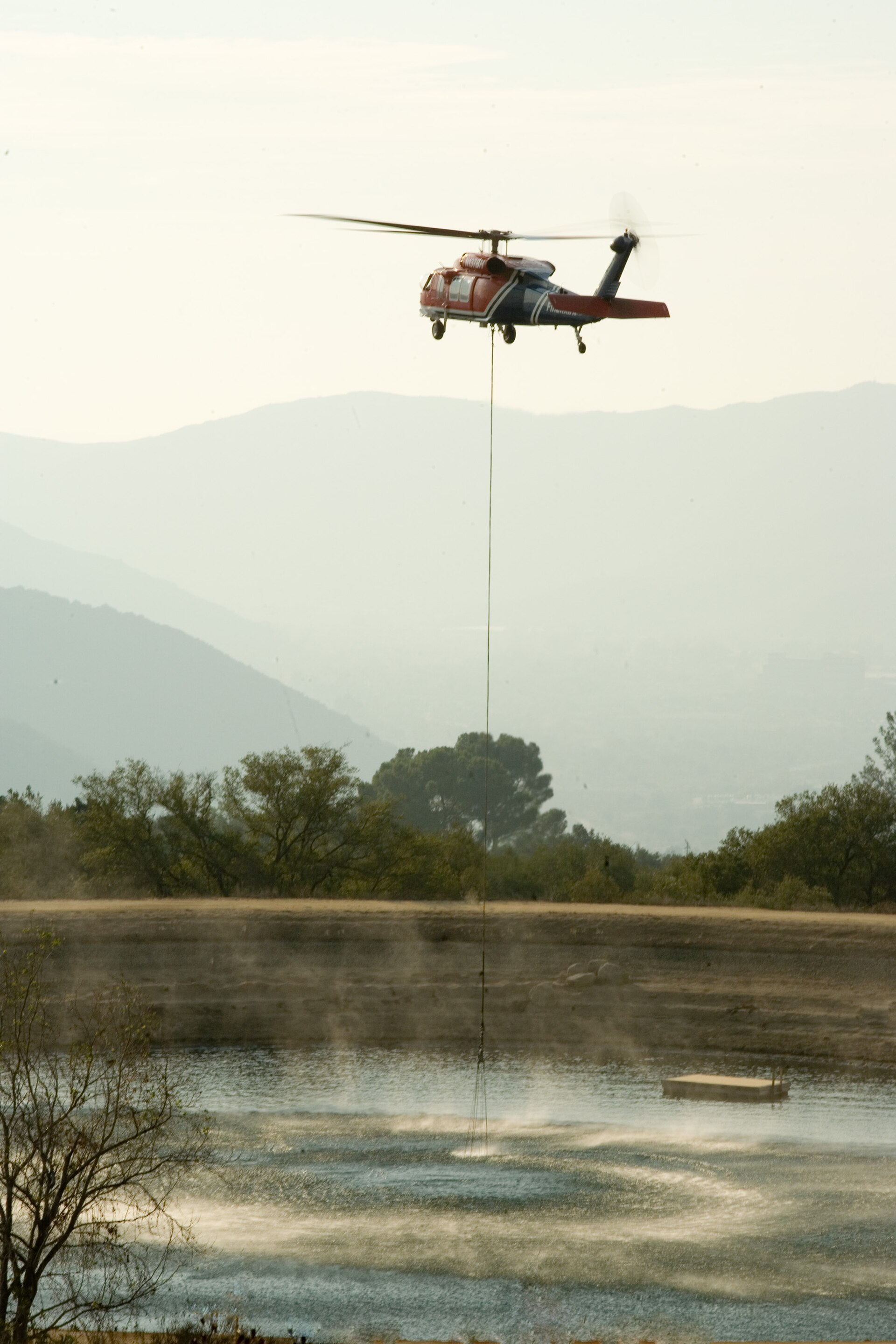 A helicopter dips a long water bucket line into a California reservoir in bright daylight