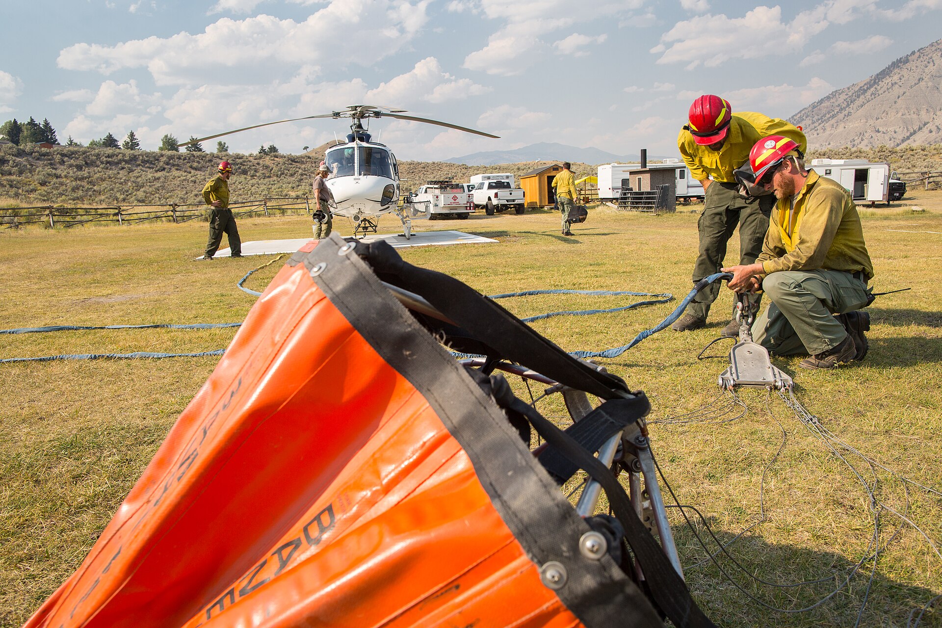 Firefighters in protective gear rig a helicopter for water bucket drops on a sunny day