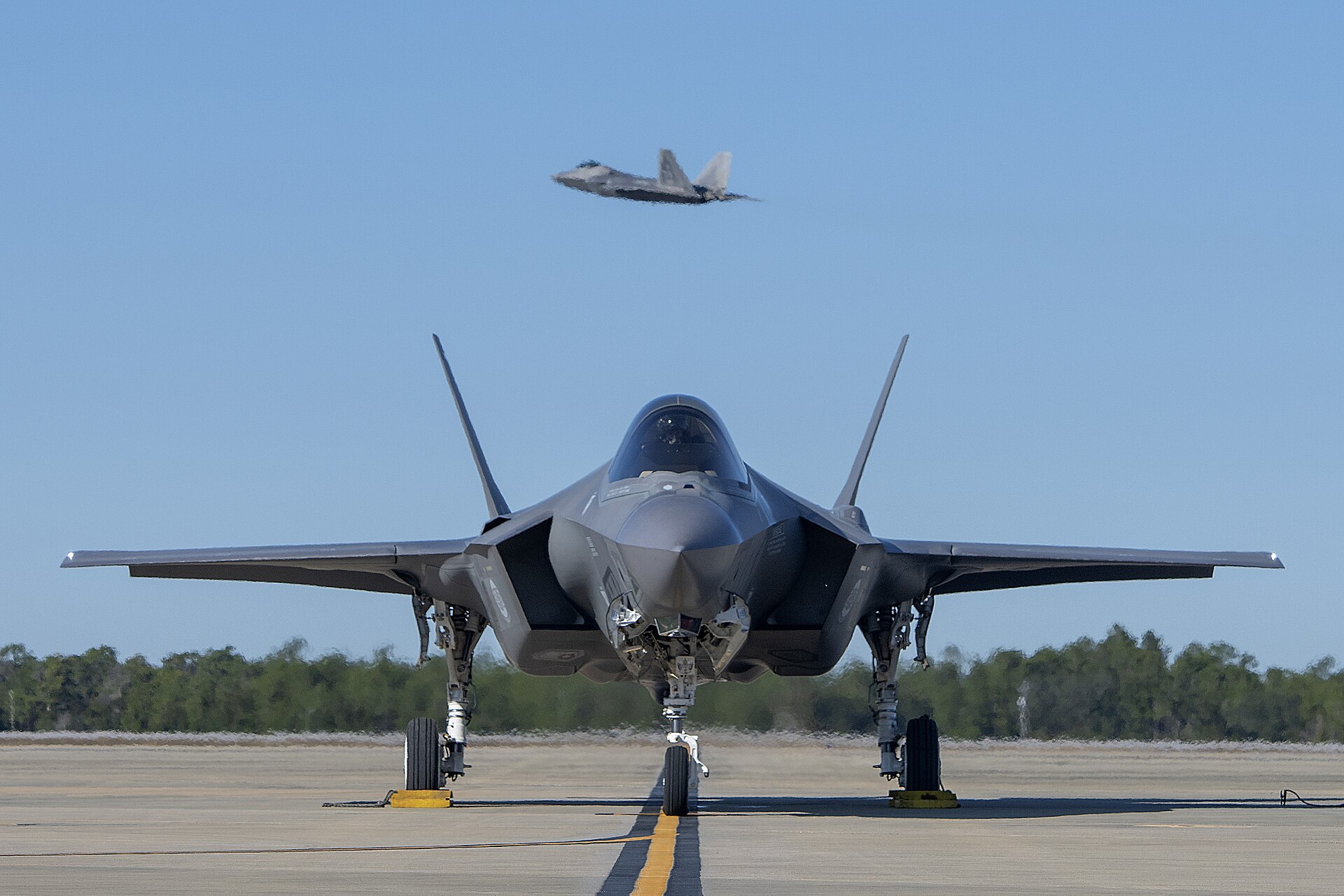 An F-35A Lightning II taxiing on a bright tarmac while an F-22 Raptor flies overhead