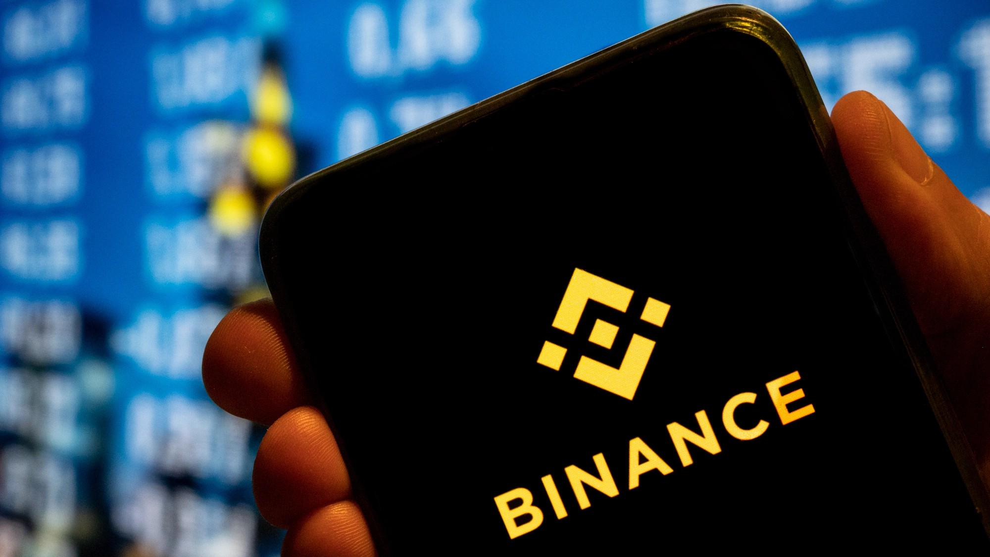 The Binance logo in gold on a smartphone screen, held in a hand against blurred blue market data.