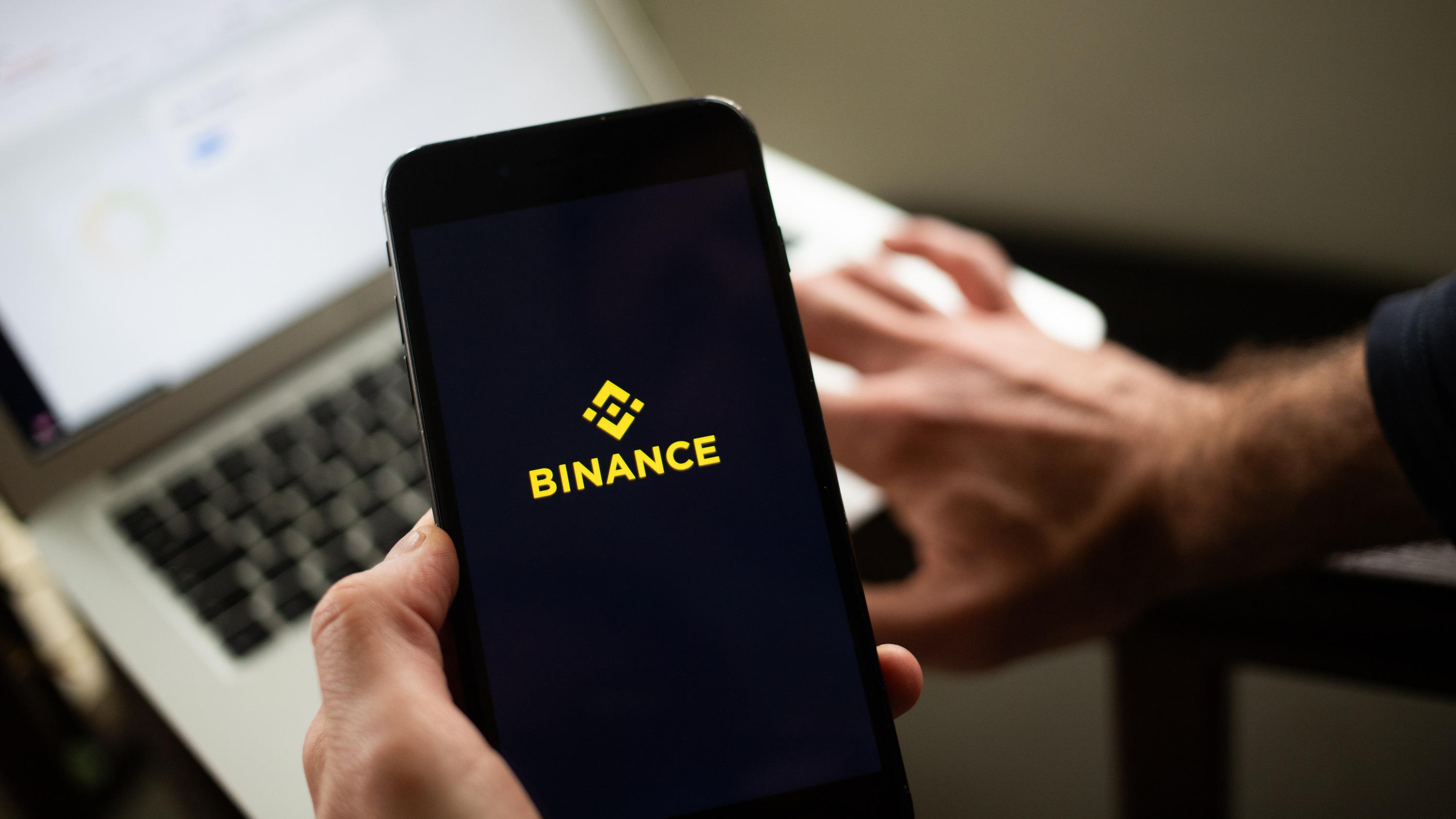 A hand holds a phone showing the Binance logo, a laptop open behind it.