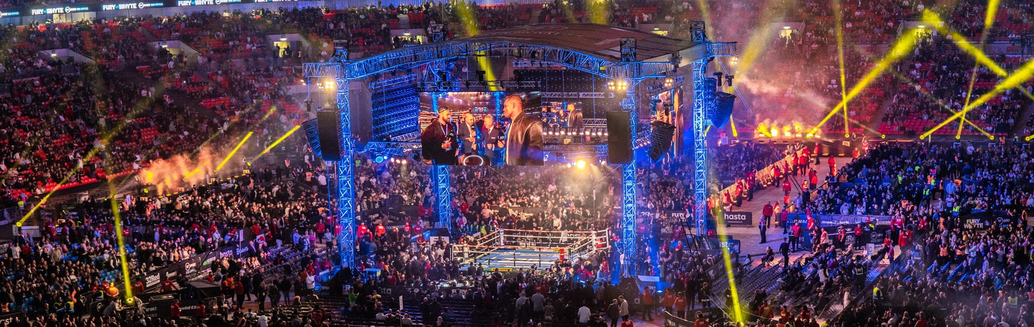 A packed stadium lit for a major boxing event — ring, big screens and crowd.