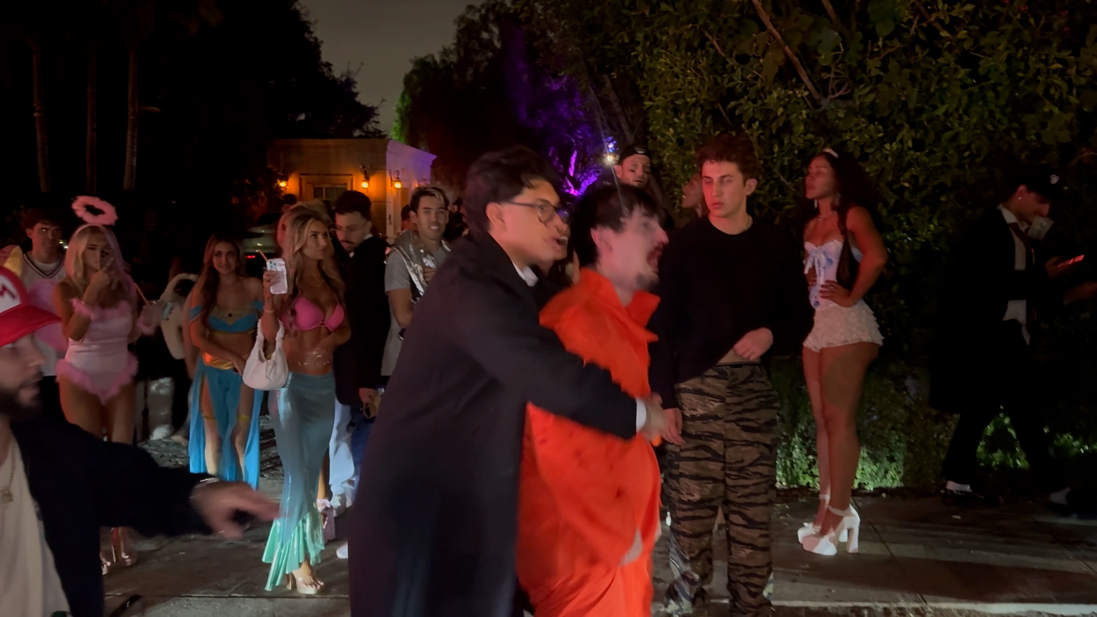 A frame from the brawl video: Hall on his feet in the orange jumpsuit, shouting while a man in a suit holds him back, costumed partygoers watching and filming.