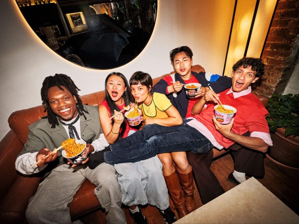 Friends laughing and eating Buldak cup noodles together on a couch