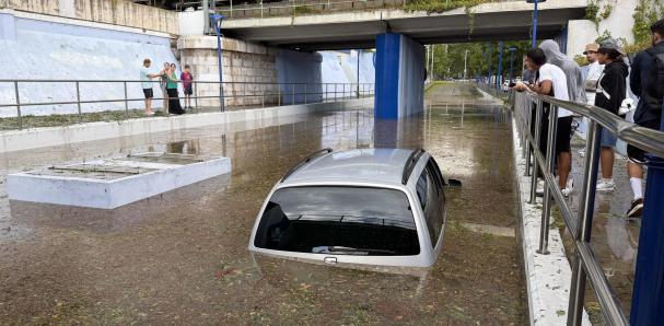 A car submerged in brown floodwater beneath a bridge in Catalonia as onlookers gather on a walkway