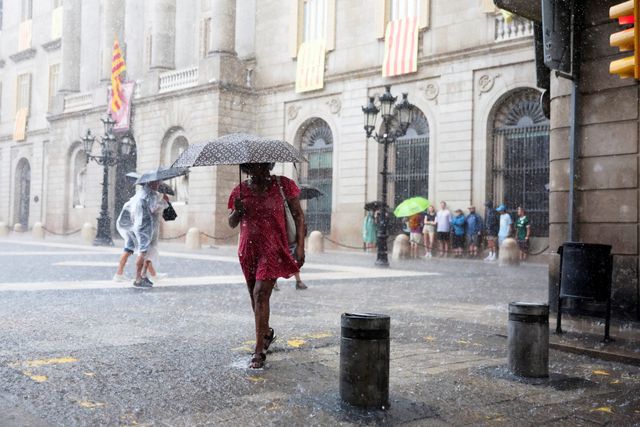 Pedestrians with umbrellas crossing a rainy Barcelona square with Catalan flags on the building behind