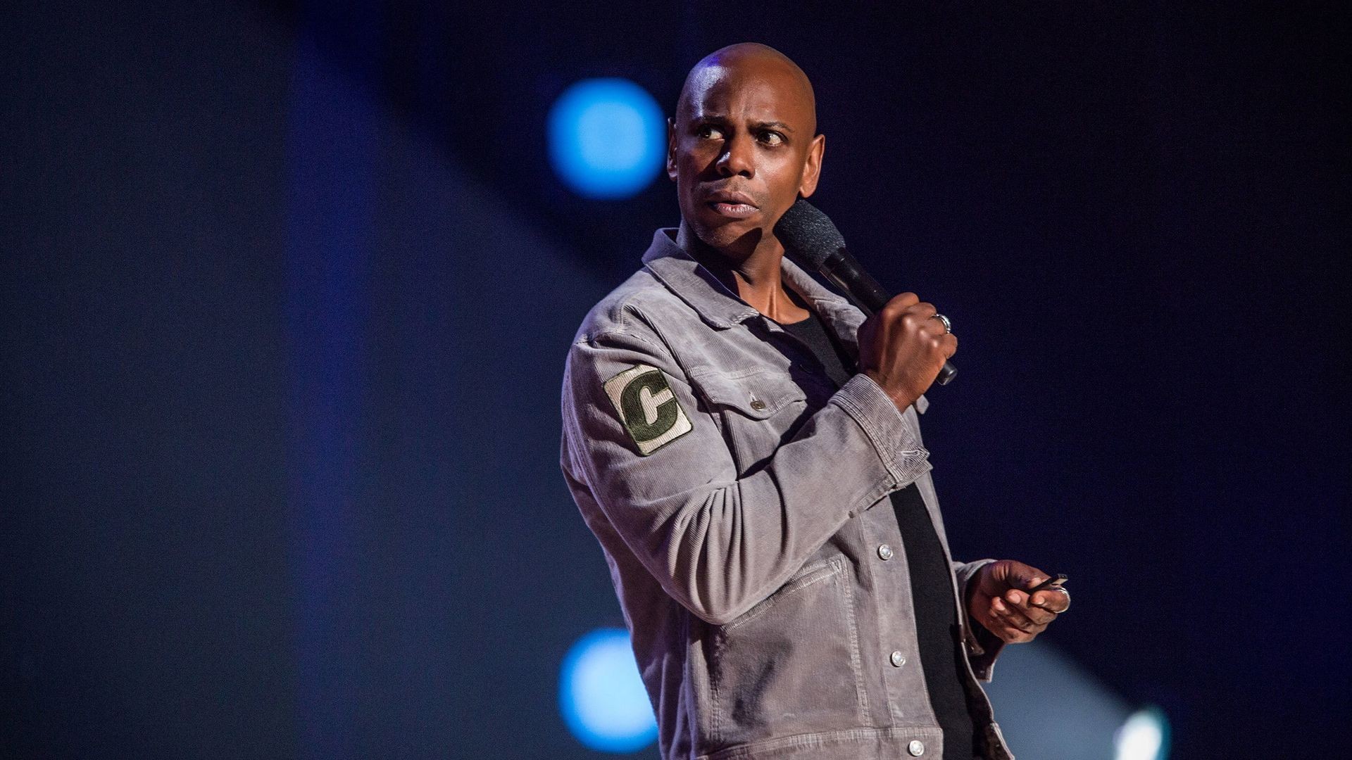 Dave Chappelle performing on stage with a microphone during his stand-up set