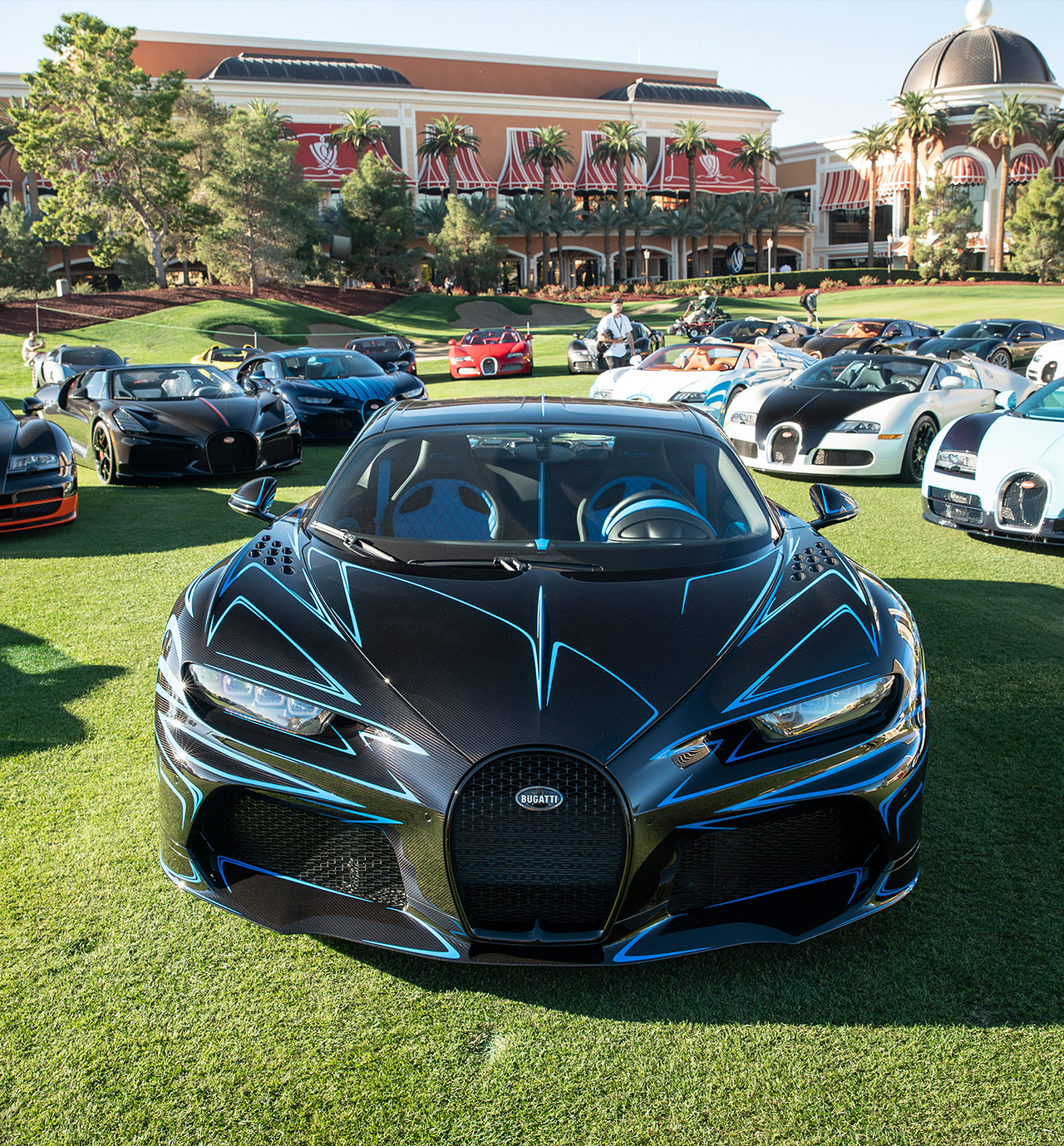 A black and blue Bugatti Chiron Super Sport nose-on on the Wynn fairways, more Bugattis behind — event file photography from a prior edition