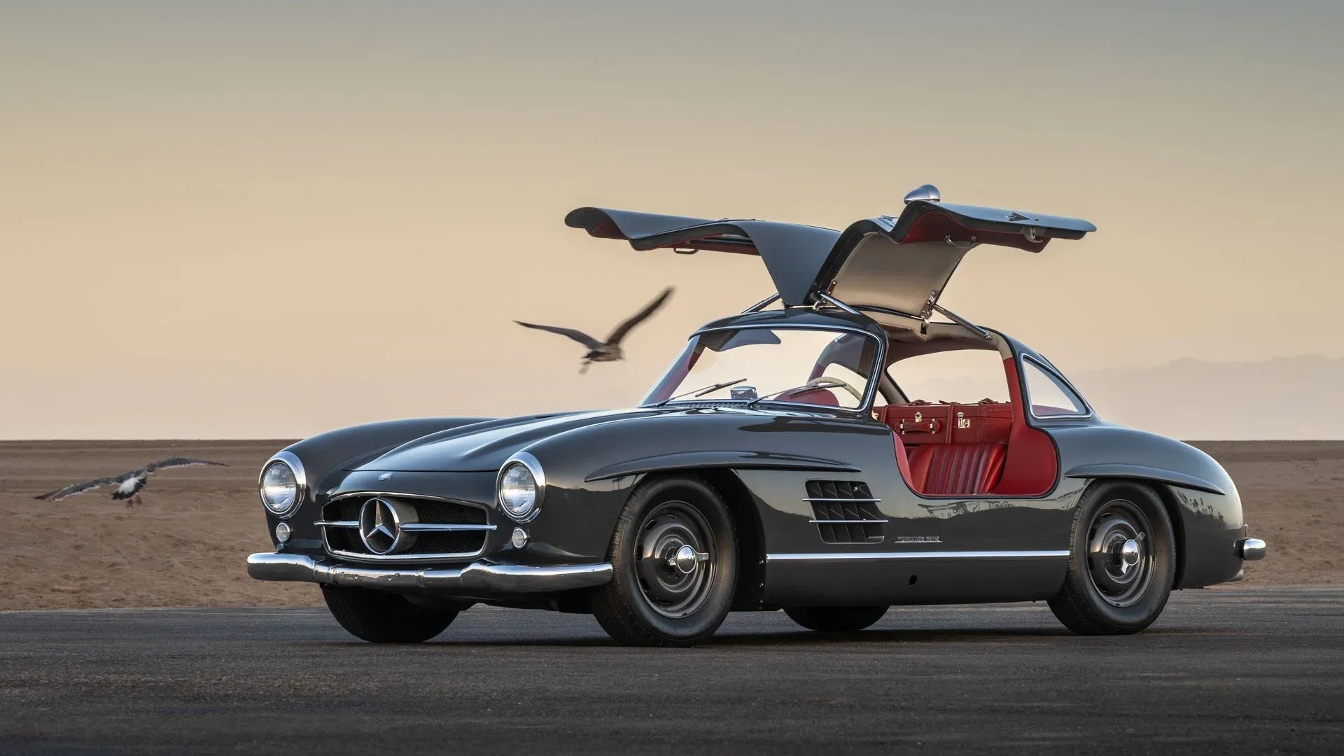 The 1955 Mercedes-Benz 300 SL Gullwing in Graphite Grey with its door raised and red interior showing — the actual car offered at the 2026 Las Vegas Auction