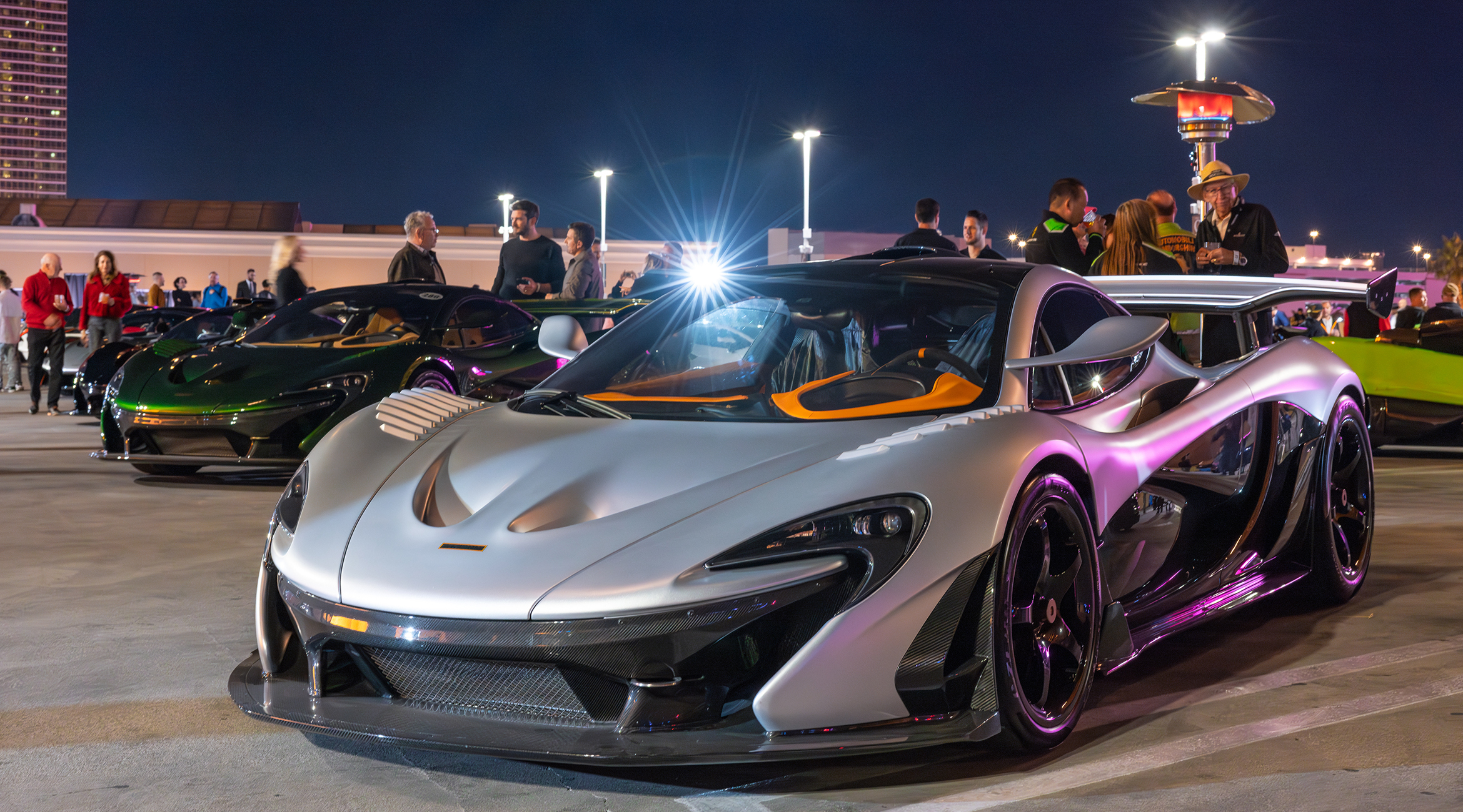 A silver McLaren P1 under the lights at Night Shift GT, the Concours at Wynn rooftop supercar night, with a green McLaren behind it and guests along the rail — event file photography from a prior edition