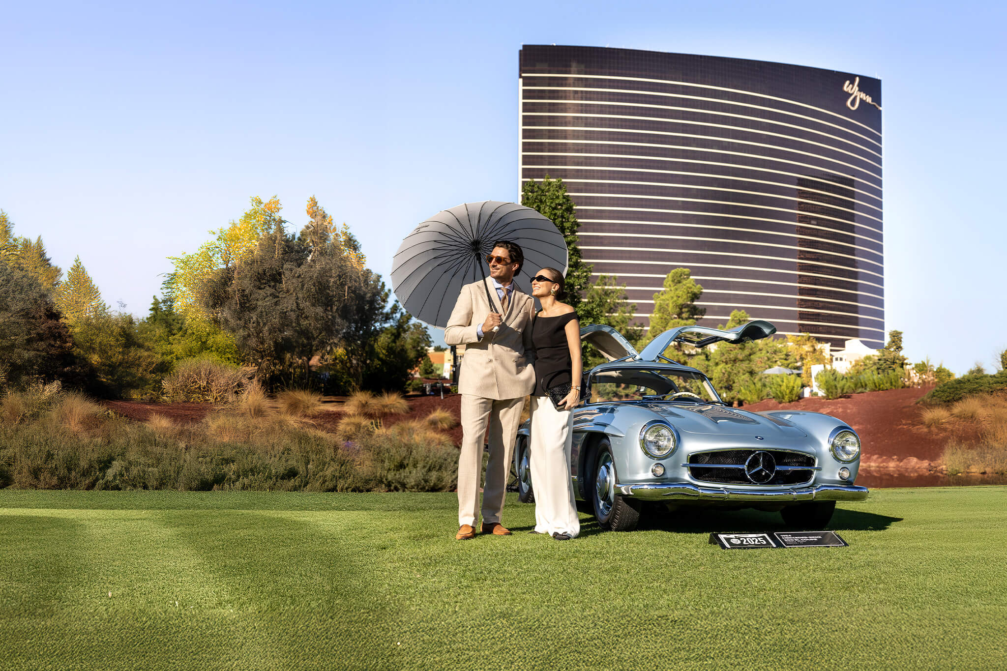 A silver Mercedes-Benz 300 SL Gullwing on the show field at last year's Concours, a couple posing beside it, the Wynn tower behind — event file photography