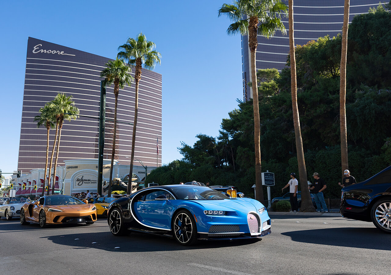 A blue Bugatti Chiron leads the Tour d'Elegance out of Wynn onto Las Vegas Boulevard, a McLaren behind it — event file photography from a prior edition