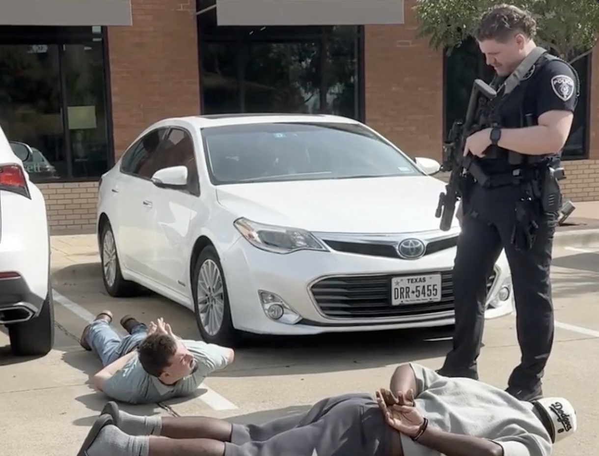 Still from the OutKick video: Dez Bryant lying face-down and handcuffed on the pavement of a Southlake, Texas parking lot while a police officer stands over the detained men holding a rifle; a second detained man lies nearby