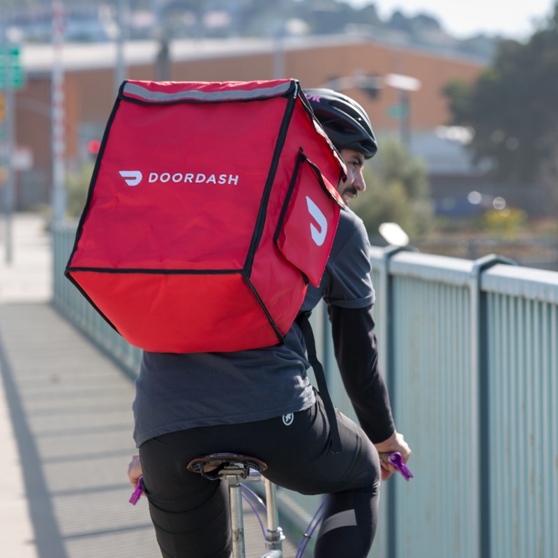 A DoorDash driver carrying a red DoorDash delivery bag on a sunny day