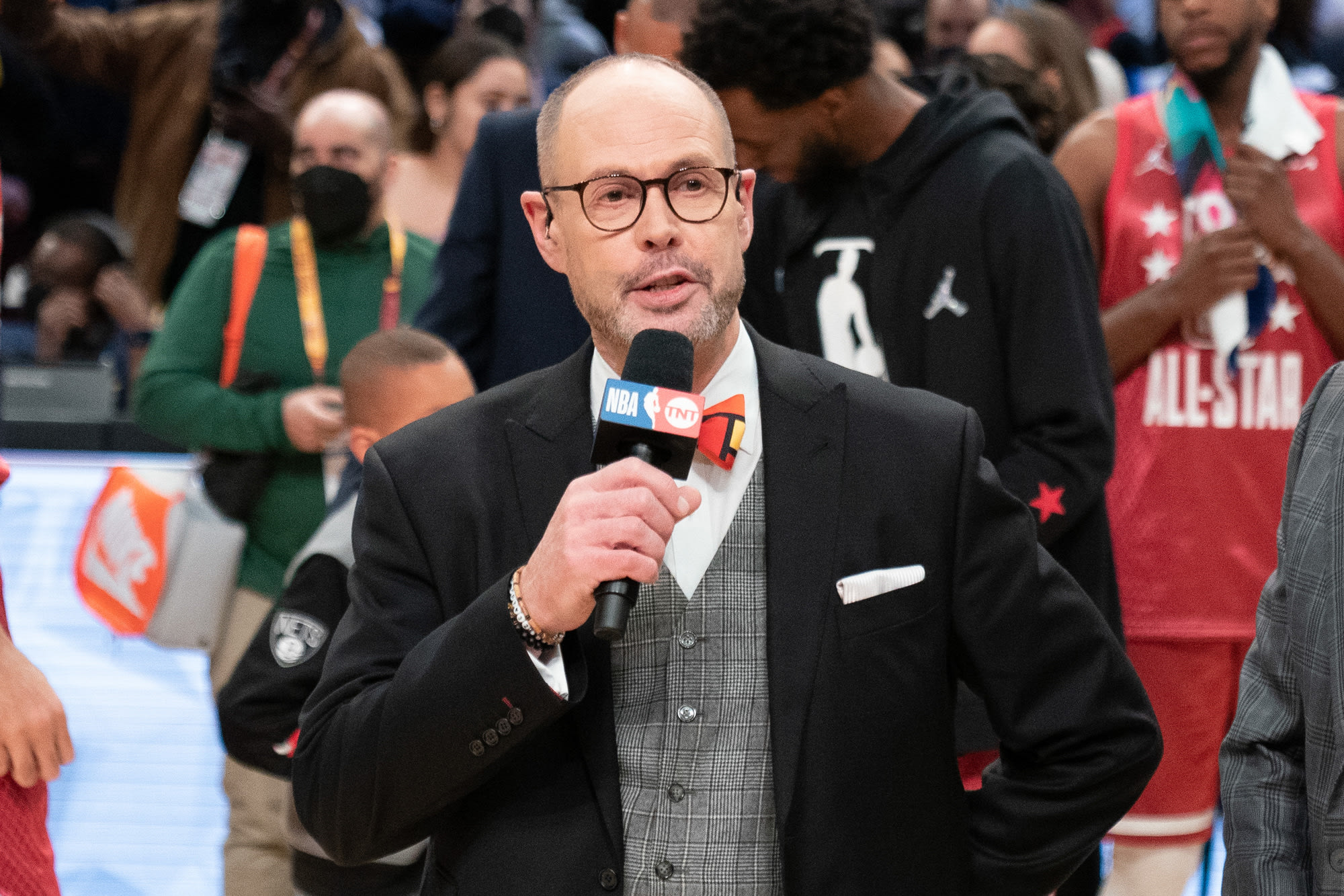 Ernie Johnson holding an NBA on TNT microphone courtside at the 2022 NBA All-Star Game in Cleveland — dark suit, plaid vest and his signature red bow tie, All-Star players behind him