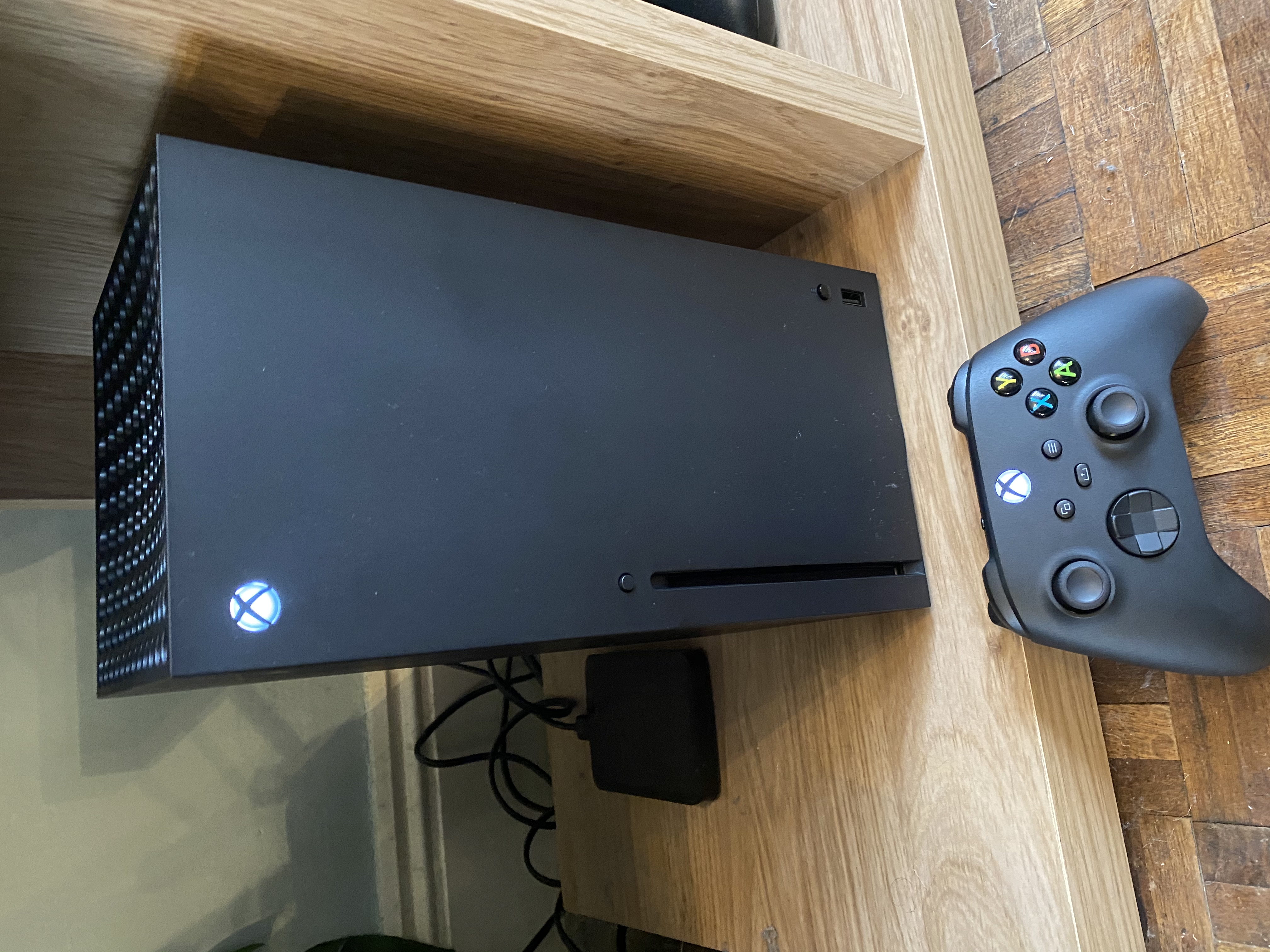 An Xbox Series X standing upright beside a wireless controller resting in a charging dock, photographed in a home gaming setup