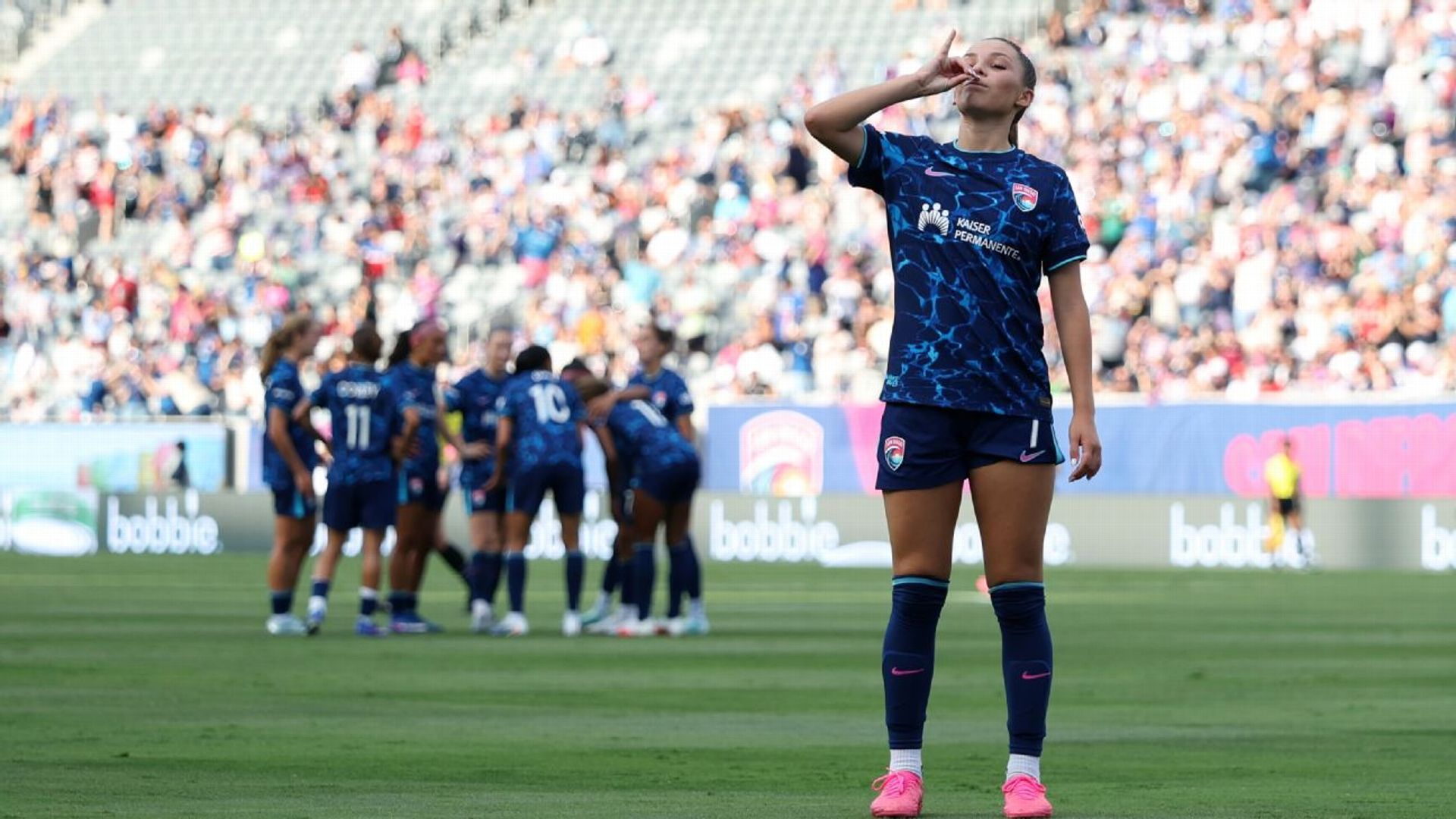 A San Diego Wave player celebrates on the pitch in front of a packed crowd, hand to her mouth, with her teammates huddled together behind her