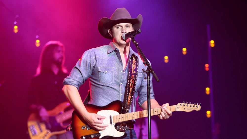 A country performer in a cowboy hat singing into a microphone on a brightly lit stage