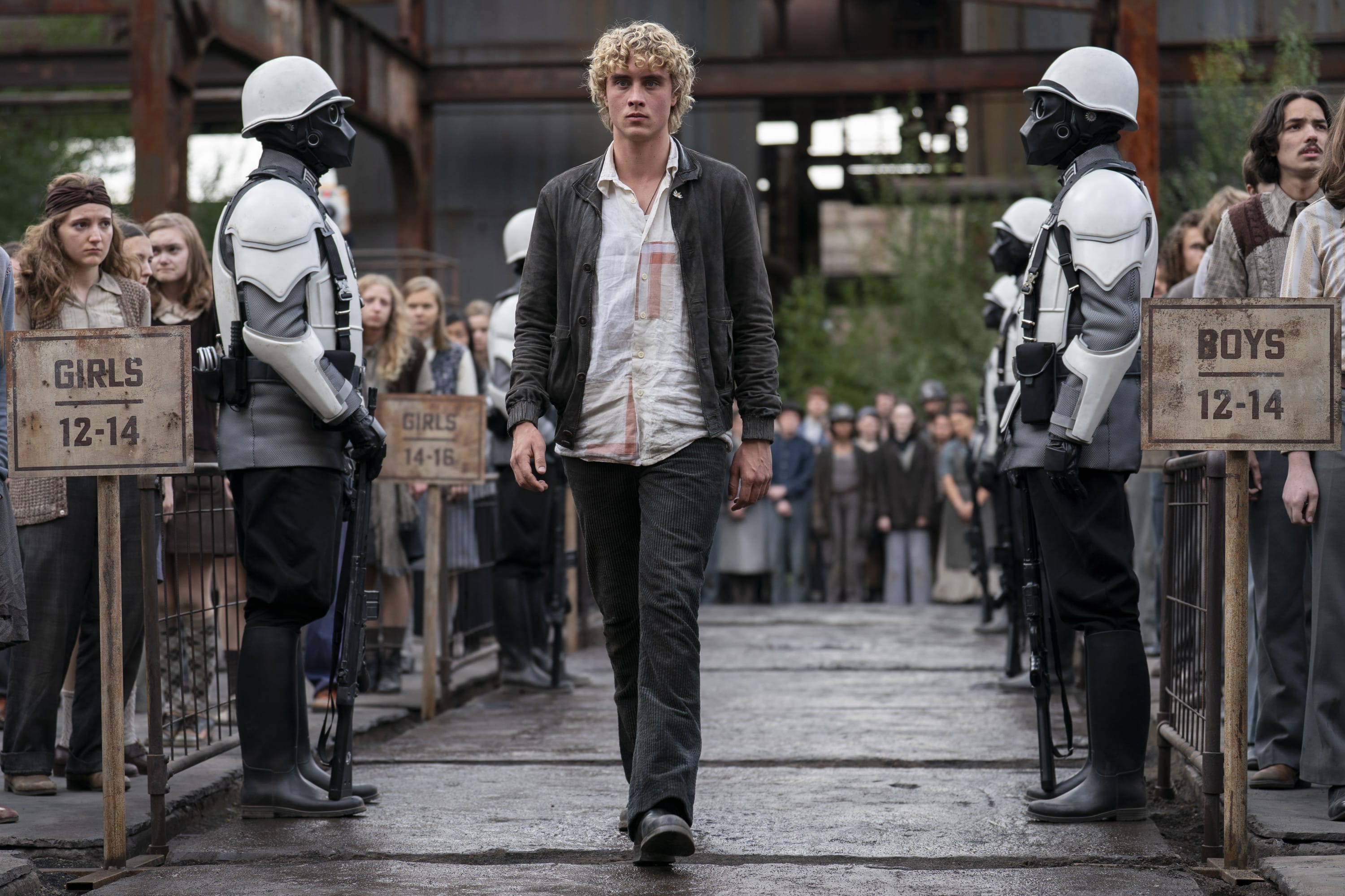 Joseph Zada as young Haymitch Abernathy walking between two Peacekeepers at the District 12 reaping in The Hunger Games: Sunrise on the Reaping, past the girls' and boys' pen signs
