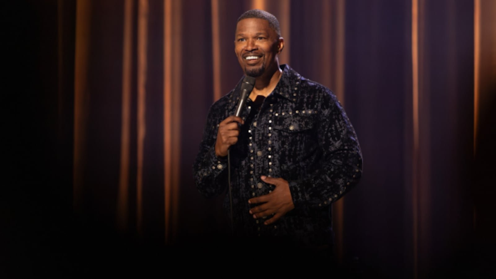 Jamie Foxx on stage holding a microphone in front of warm curtains, from his Hulu stand-up special Take Care of Yourself