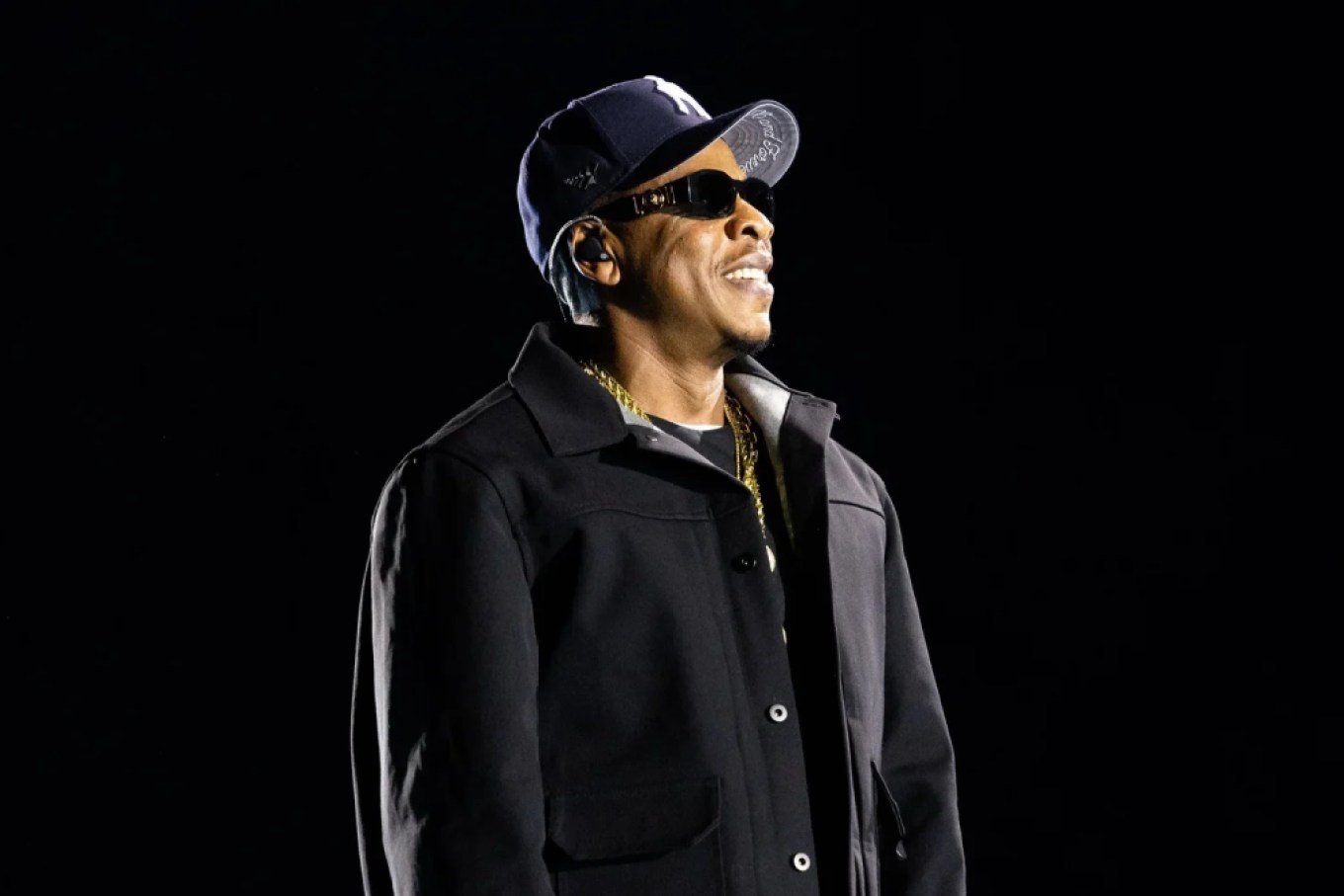 Jay-Z on stage at SoFi Stadium during his JAY-Z30 stand, in profile — black Yankees cap, sunglasses, black jacket and gold chains against a black backdrop