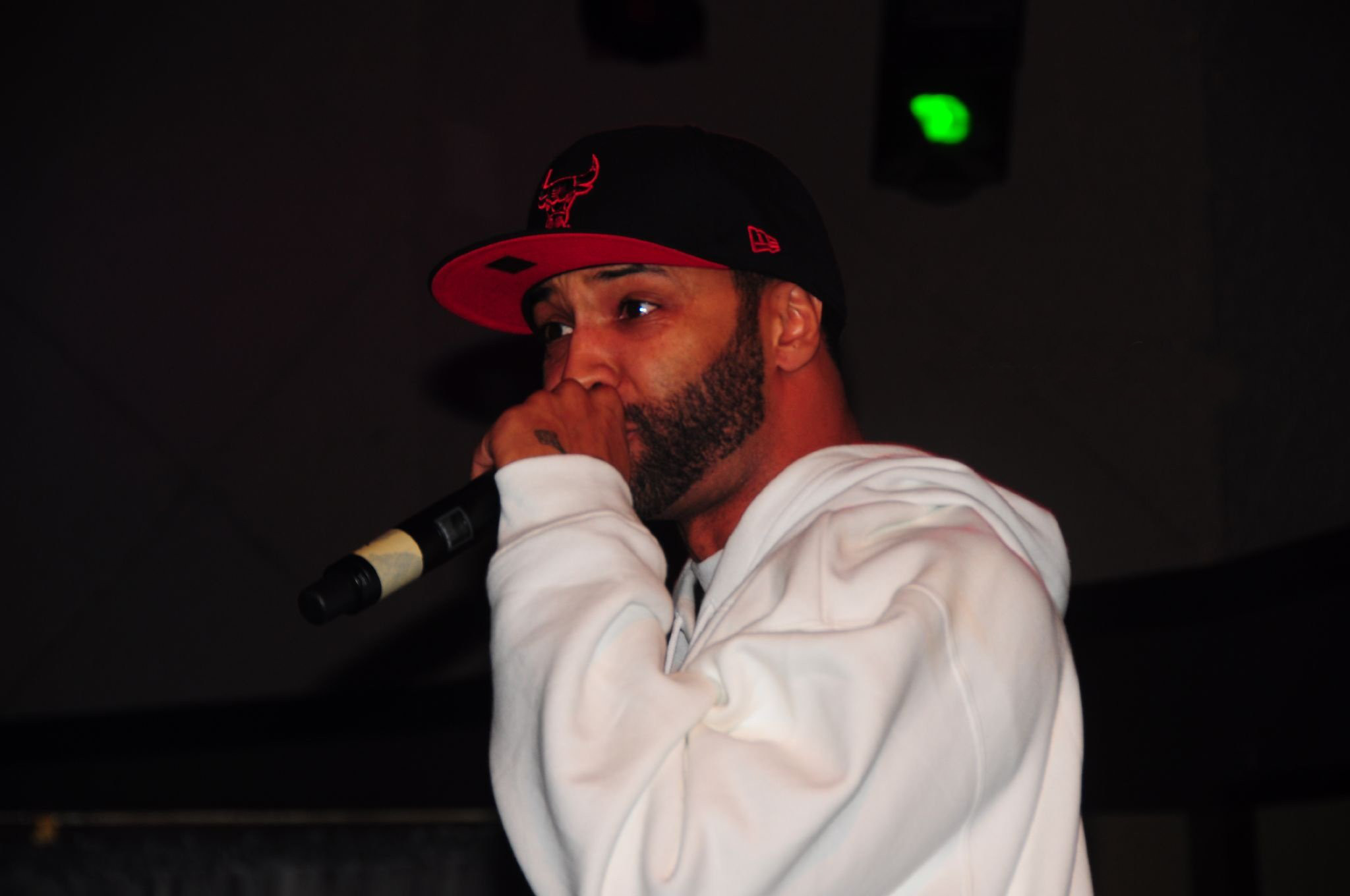 Joe Budden performing on stage in a white hoodie and black-and-red cap, microphone in hand