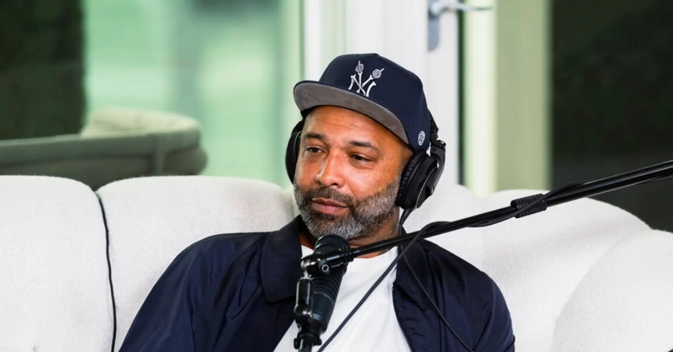 Joe Budden on his podcast couch wearing headphones and a Yankees cap, speaking into a studio microphone