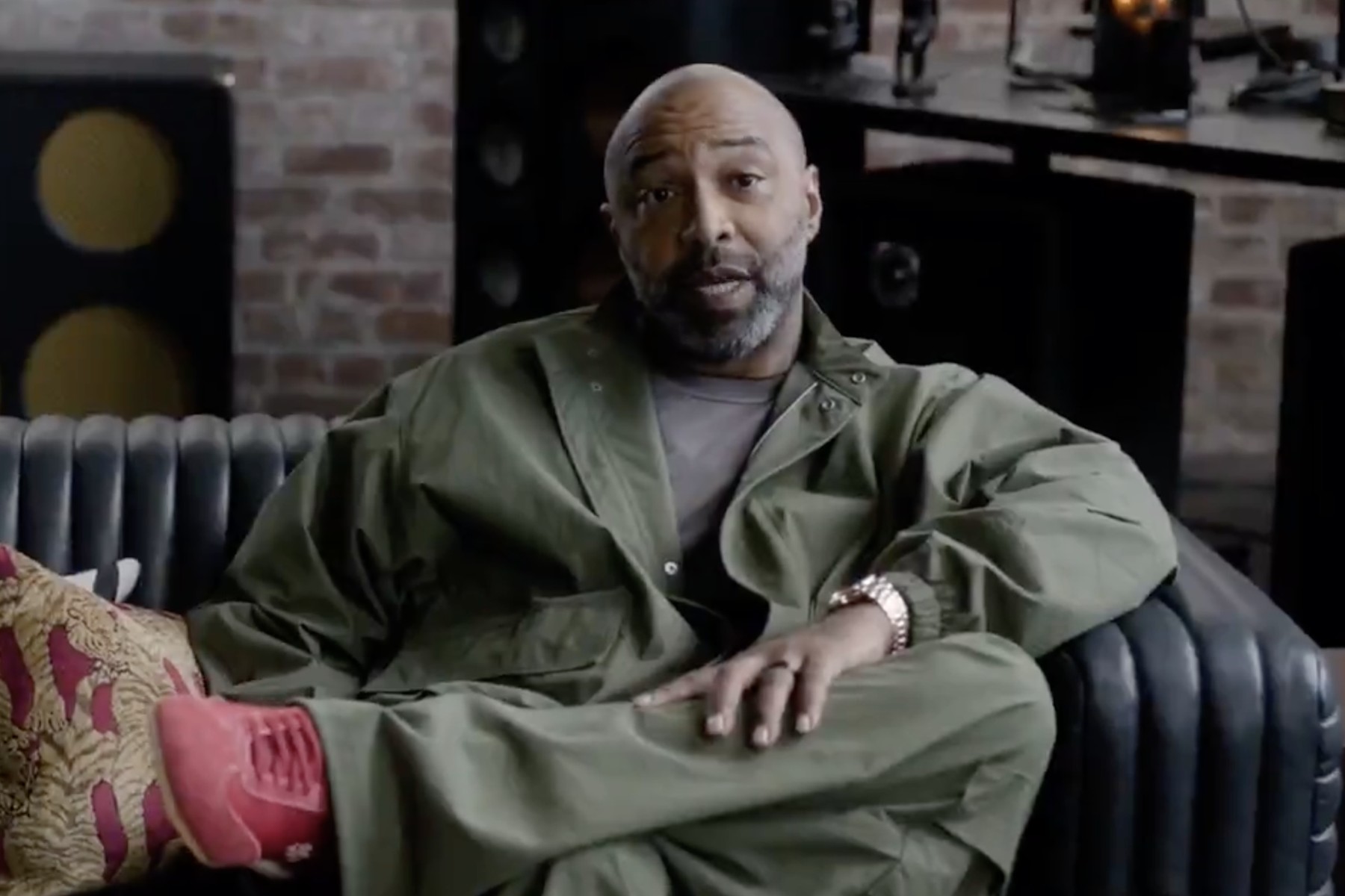 Joe Budden as Big Switty, seated on a couch in an olive outfit during his Law & Order appearance