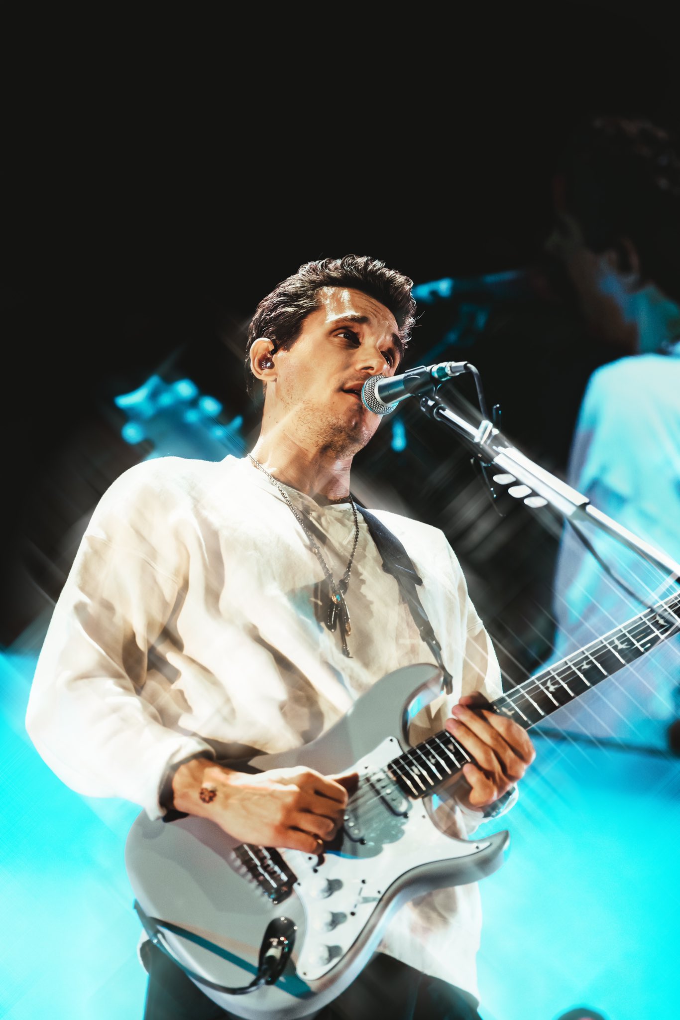 John Mayer performing live in 2019, singing into a microphone and playing a silver electric guitar in a white long-sleeve shirt