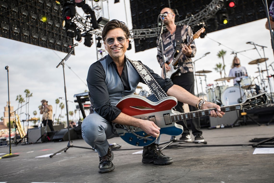 John Stamos performing on stage with The Beach Boys, drums in hand.