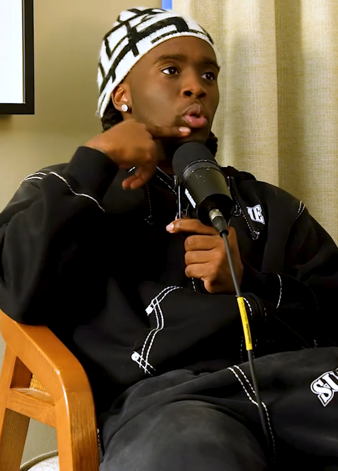 Kai Cenat seated mid-interview with a microphone, wearing a black-and-white beanie and black hoodie, during a 2023 Million Dollaz Worth of Game interview