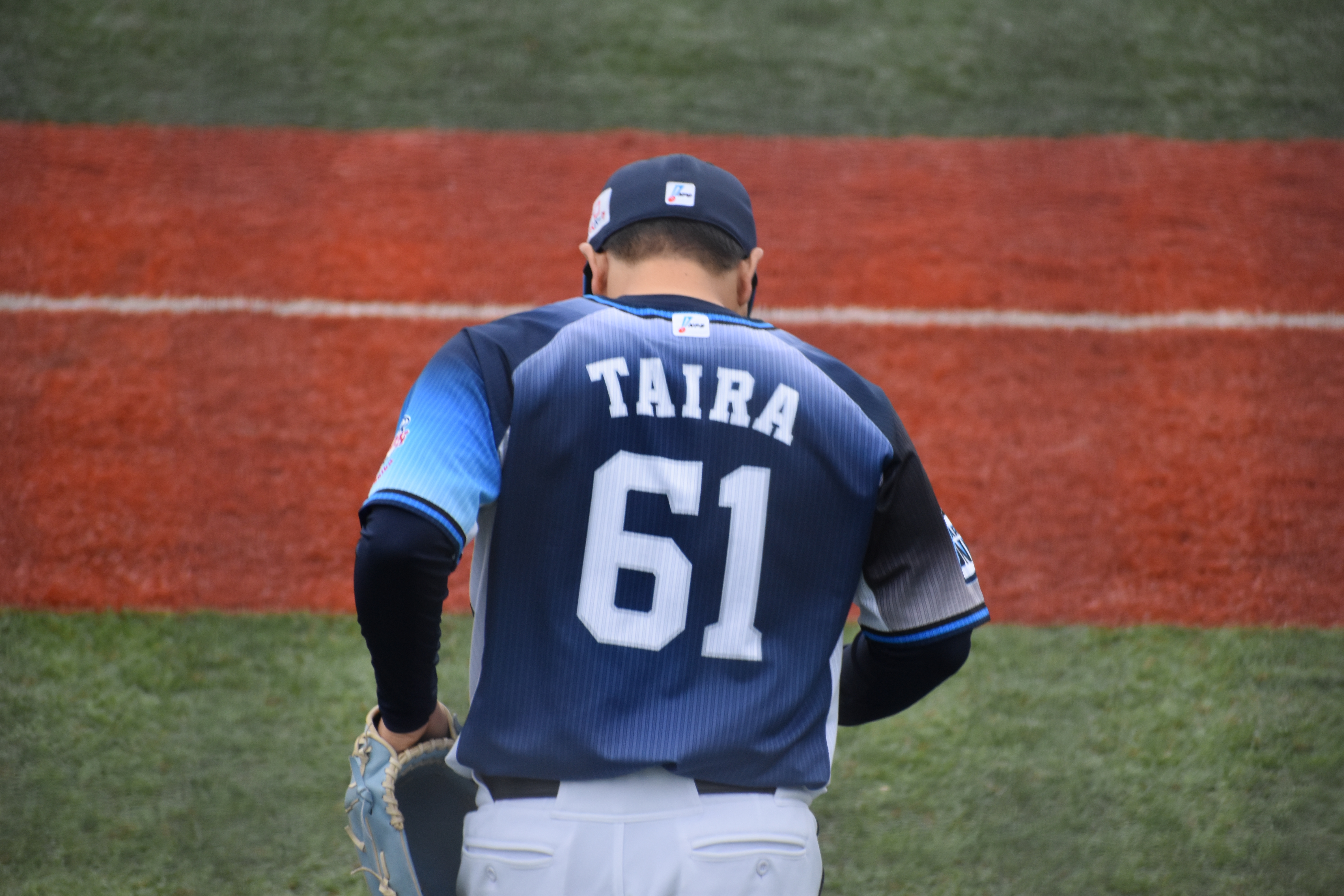 Kaima Taira from behind in his Saitama Seibu Lions jersey, his name and No. 61 across his back, glove in hand on the field at Kamagaya Stadium