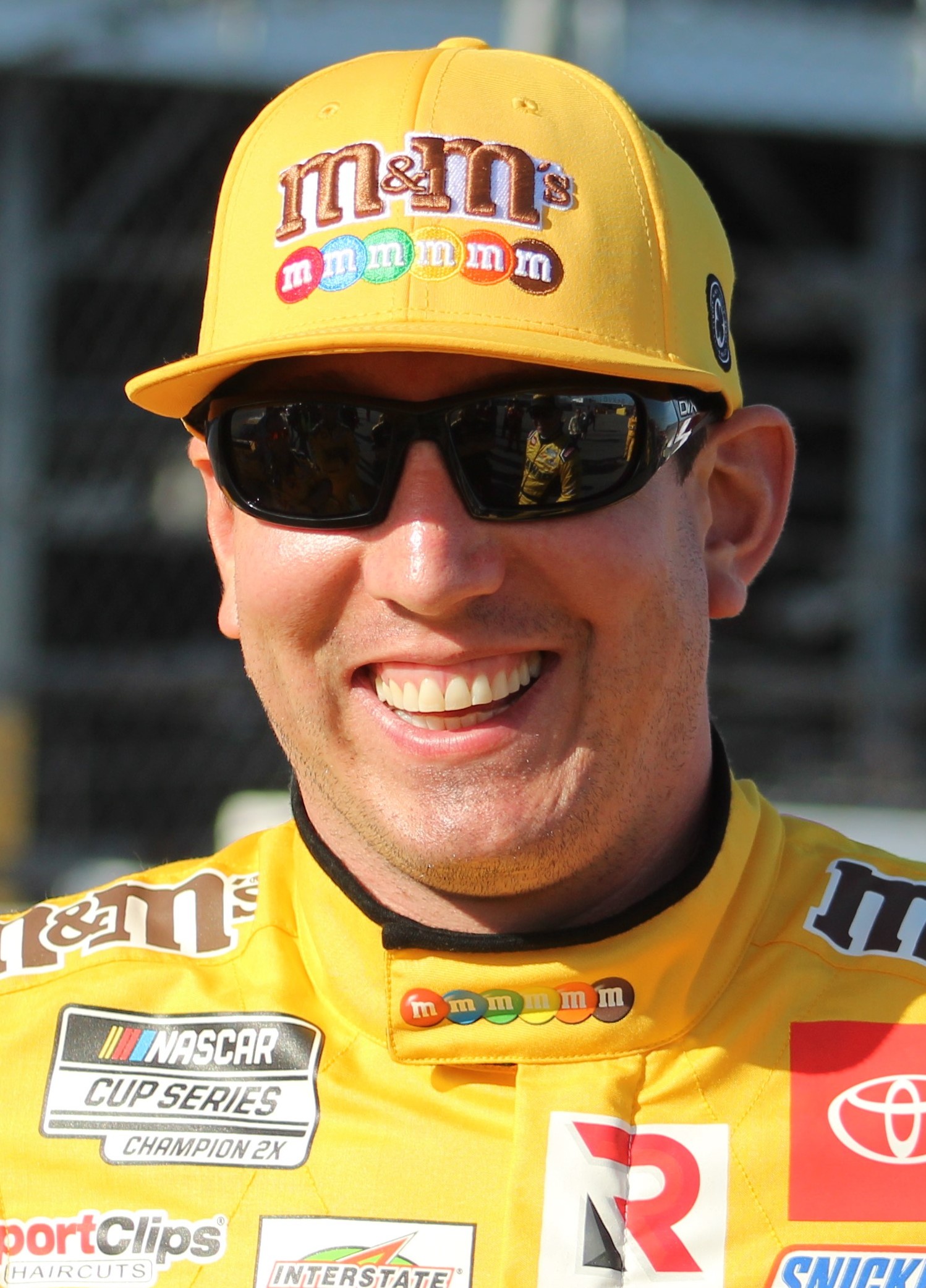 Kyle Busch smiling in his yellow M&M's firesuit and cap at a NASCAR race