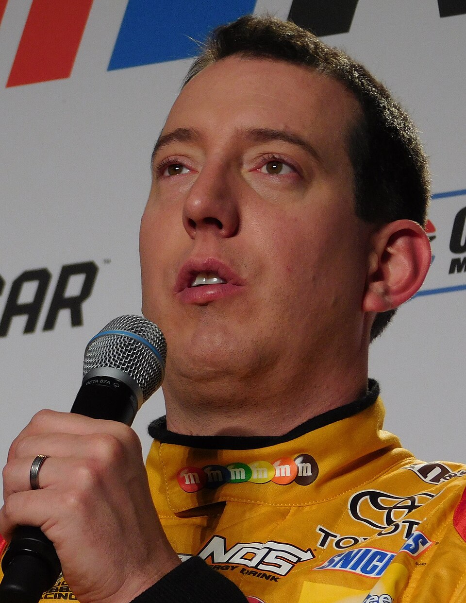Kyle Busch speaking into a microphone in his firesuit during the 2017 NASCAR Media Tour