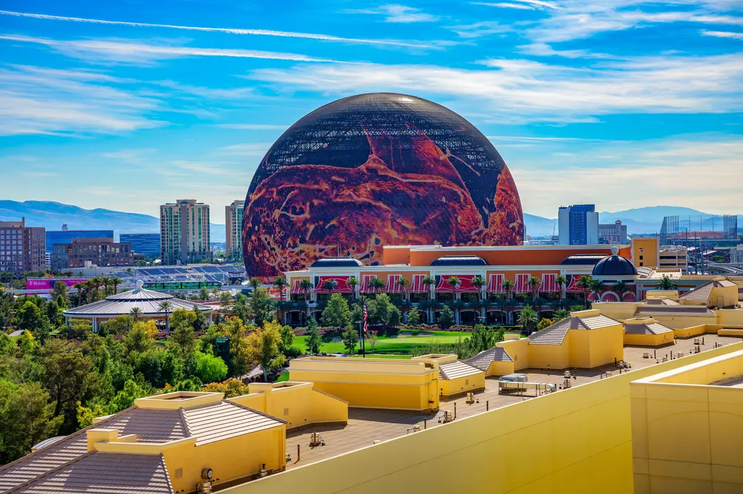The Sphere in Las Vegas displaying red and orange visuals on its exterior LED screen on a bright day, with the Wynn in the foreground
