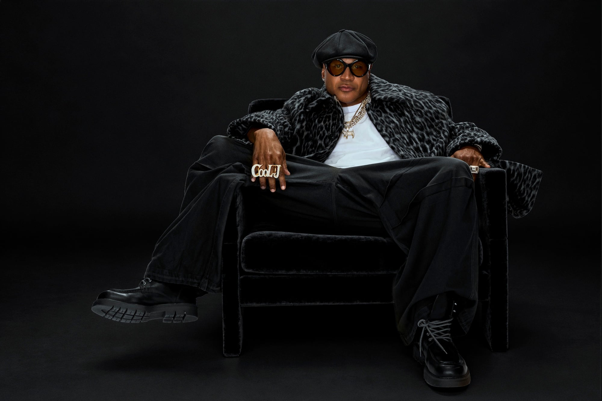 LL Cool J in the Rock the Bells: The Residency announcement portrait — seated in a black cap, sunglasses and a leopard-print coat, wearing his 'Cool J' ring