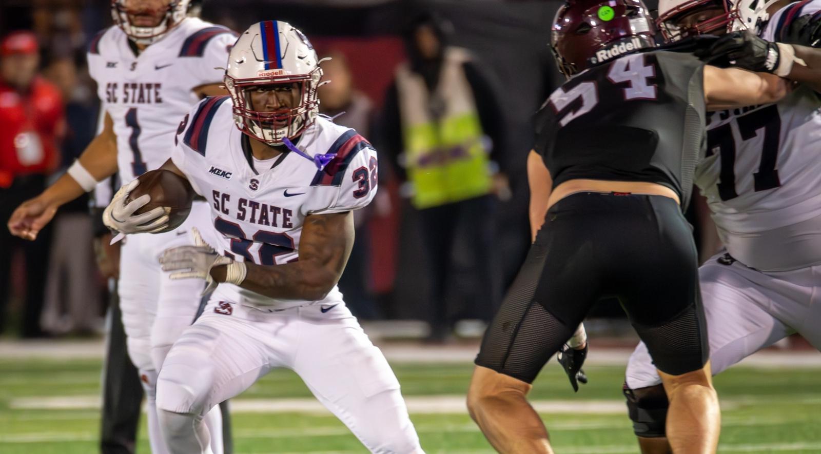 A South Carolina State ball carrier (No. 32, white SC State uniform, MEAC patch) runs with the football against a defender in black (No. 54) under the lights.