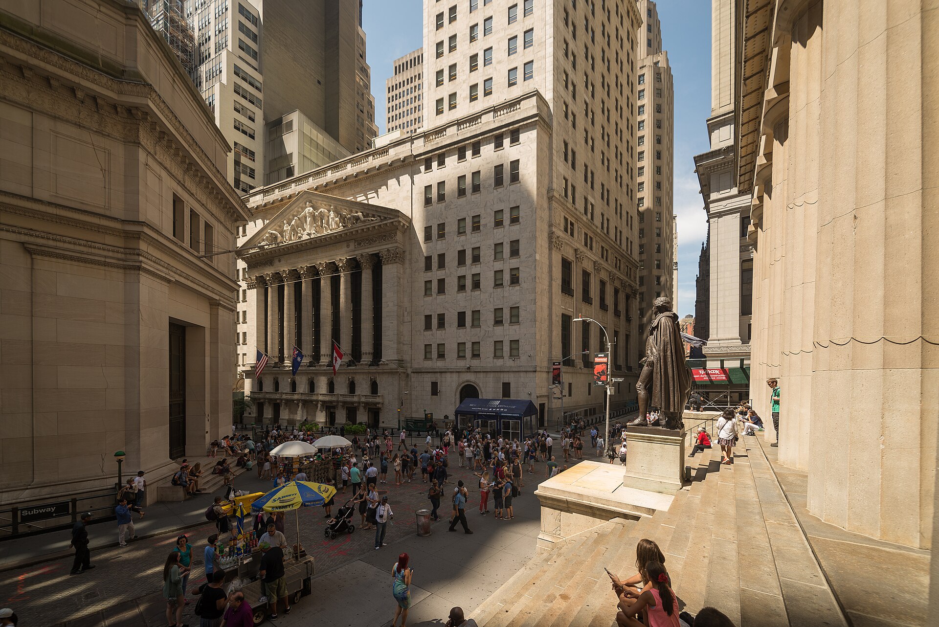 The New York Stock Exchange on Wall Street on a bright daytime, with crowds gathered outside the landmark facade.