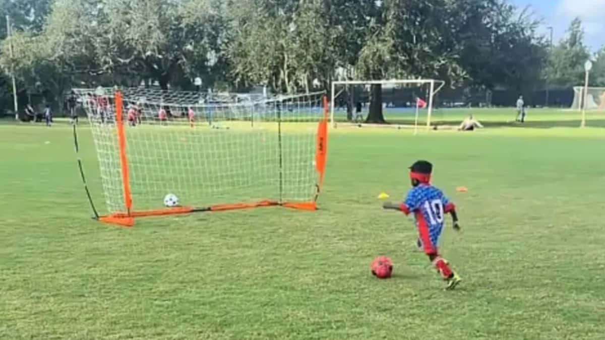 Three-year-old Nevada in his blue and red No. 10 kit striking a red soccer ball toward an orange practice goal on a grass field in Florida