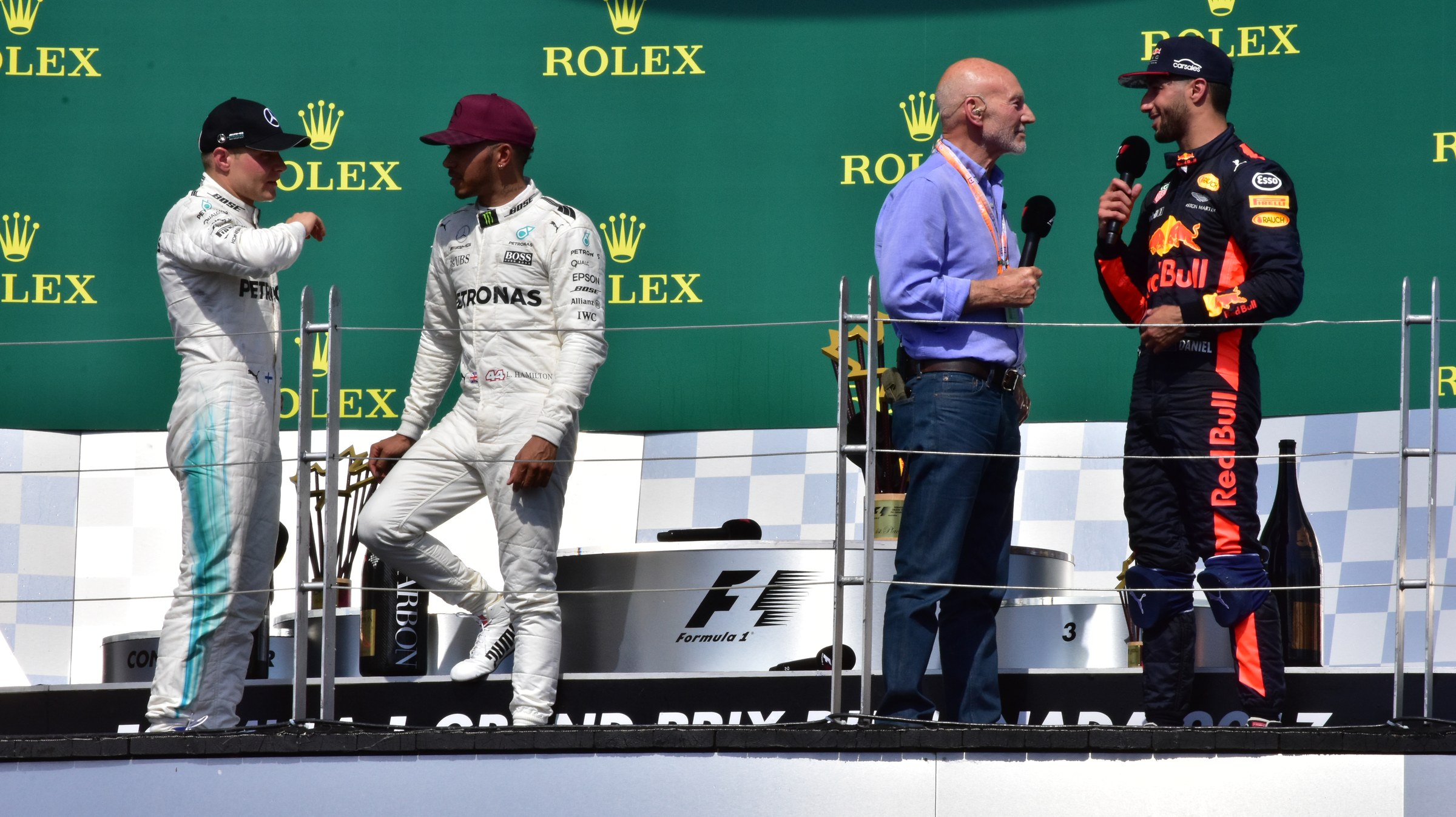 Patrick Stewart holding a microphone on the podium at the 2017 Canadian Grand Prix, interviewing the finishers alongside Lewis Hamilton, Valtteri Bottas and Daniel Ricciardo