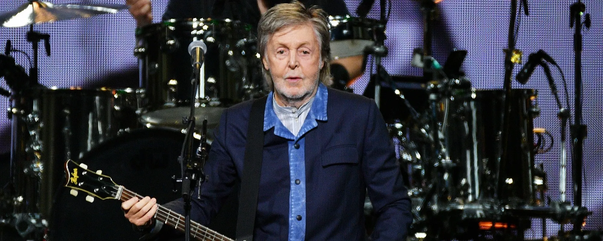 Paul McCartney performing on stage with his bass guitar, his band's drum kit behind him against a lit backdrop