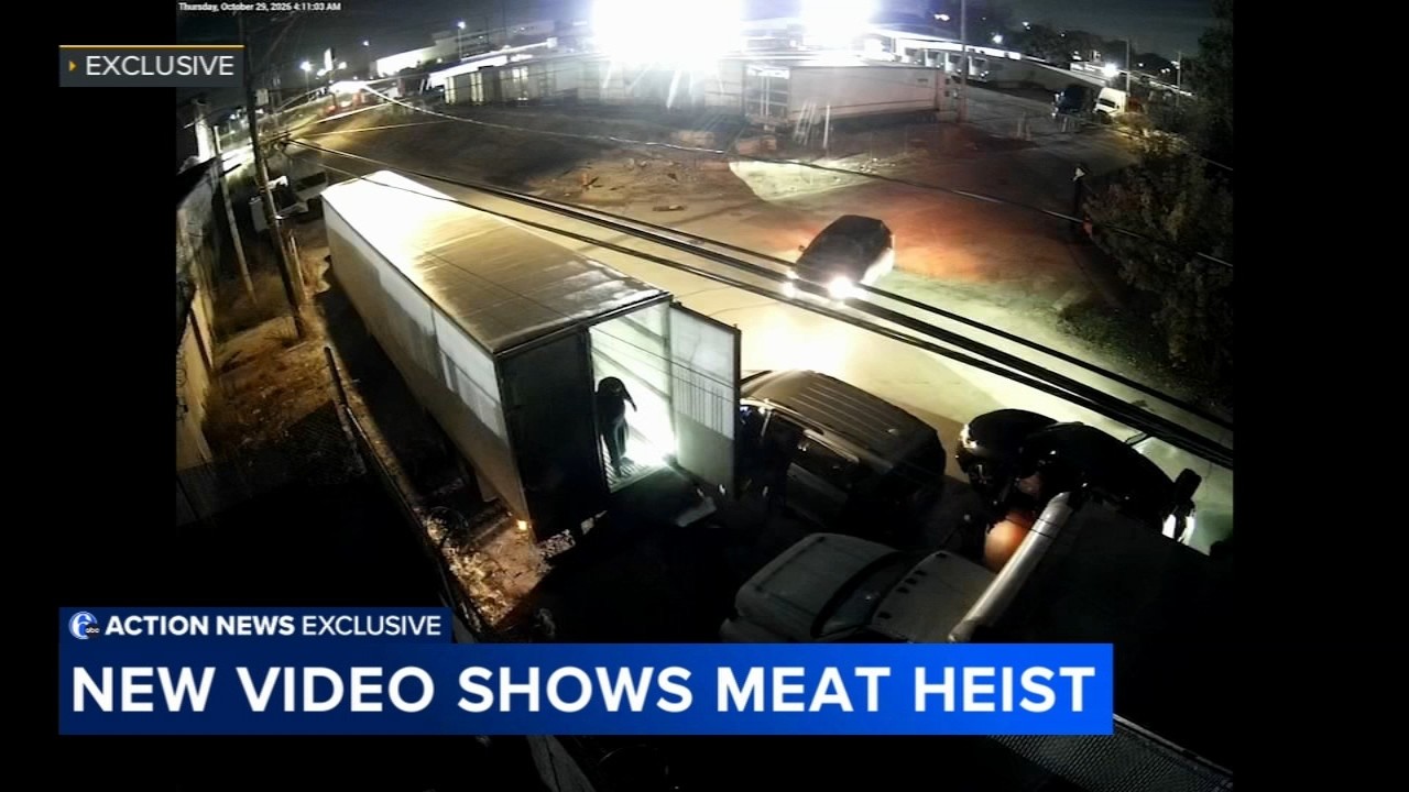 Surveillance frame timestamped Thursday, October 29, 2026, 4:11 a.m.: a figure stands in the lit doorway of an opened trailer while vehicles wait alongside during the Devereaux Street beef heist.