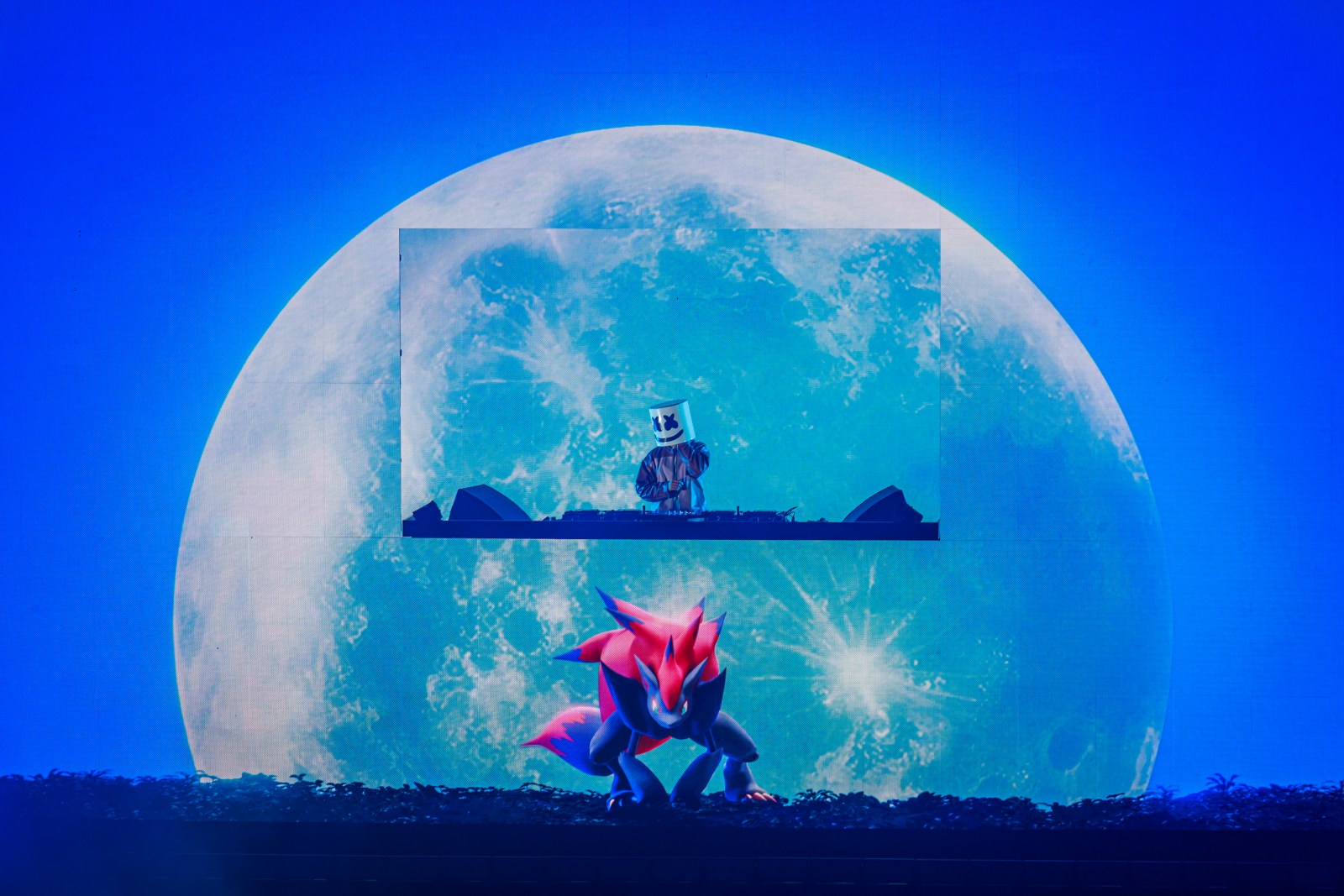 Marshmello at the DJ decks during Pokémon Night Out at the Intuit Dome — centred in a video-wall inset against a giant full-moon visual, with Zoroark on the stage screen below him and deep blue arena light throughout