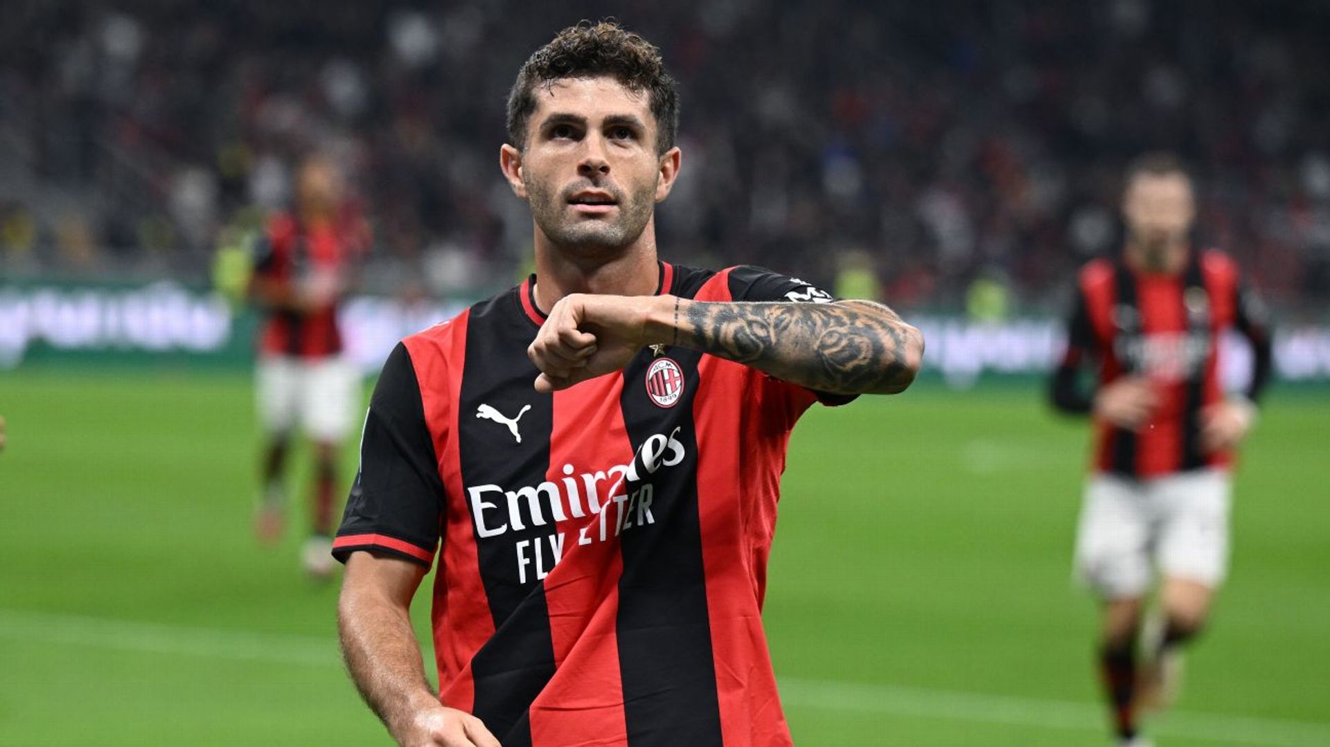 Christian Pulisic in AC Milan's red-and-black home kit, fist raised in celebration, teammates blurred behind him at San Siro